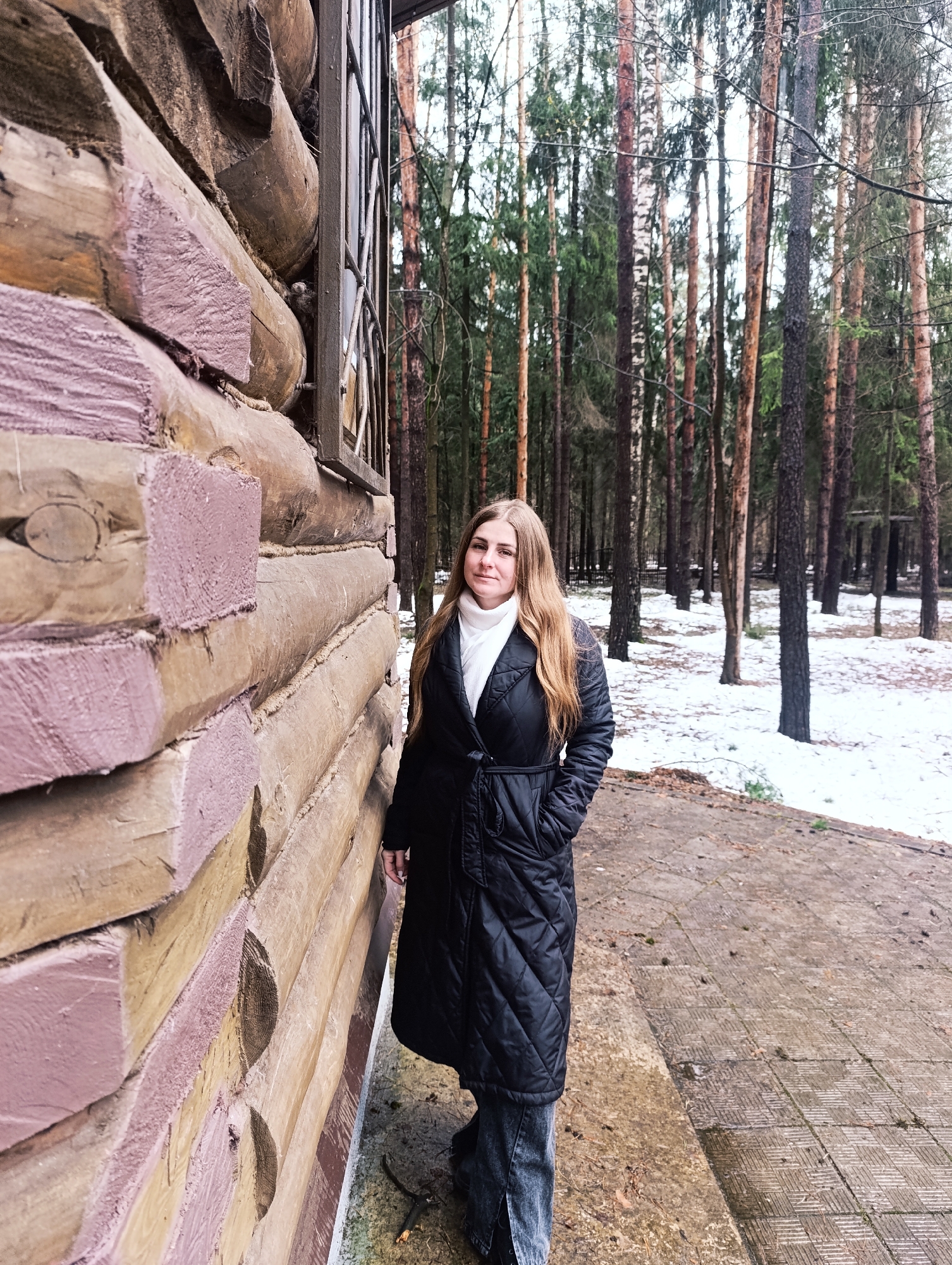 A young woman stands outdoors beside a rustic log building, backed by a dense forest, likely in Chernogolovka, Russia. She has long, light brown hair and is wearing a white turtleneck sweater, a black quilted long coat, and dark pants, with her hands in her pockets. She looks directly at the viewer with a neutral expression.
To her left, the log wall of a building is prominent, constructed from large, rough-hewn logs. The cut ends and some sides of the logs are painted a muted pinkish-purple, contrasting with the natural wood color of other logs. Rope caulking is visible between the log layers. A dark metal grate or fence extends upwards along the building's side, beyond the logs.
The ground in the immediate foreground is covered with light grey square paving stones, with some debris. Beyond this, the forest floor is visible with patches of snow and fallen leaves. The background features a thick stand of tall, slender trees, predominantly evergreens such as pines and spruces, interspersed with lighter-barked deciduous trees like birches. The presence of snow indicates cold weather, possibly late winter or early spring. The lighting is soft and diffused, suggesting an overcast daytime sky. No text is visible in the image.