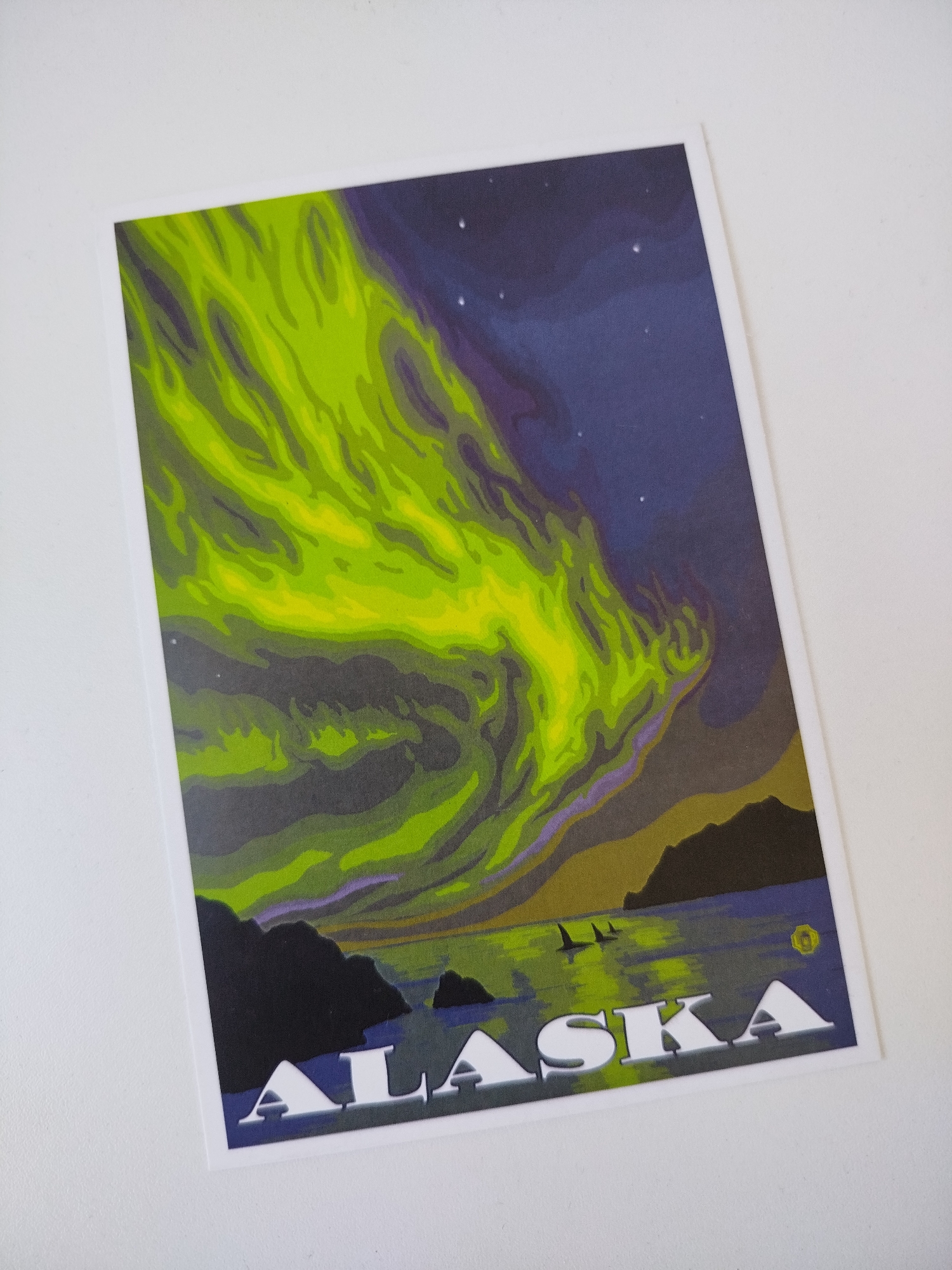 The image is a postcard of Alaska. The postcard depicts a landscape with an aurora borealis in the sky. There are land masses, water, and two whale silhouettes in the image. The word "ALASKA" is printed in large, white letters at the bottom of the card. The photo is taken on a white surface, and the postcard is angled slightly. There is no indication that this photo was taken in Varna, Bulgaria.
