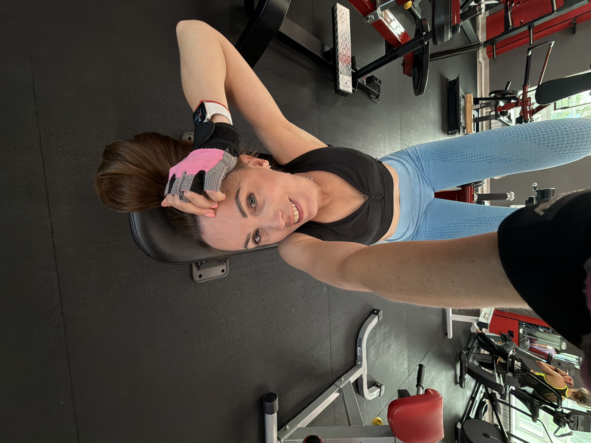 The image depicts a woman upside down on a gym bench, taking a selfie. She is wearing a black sports bra and light blue leggings. Her hair is brown and falls around her face. She is wearing workout gloves on her hands, one with pink accents. The gym environment includes various weight machines and equipment, with a dark rubberized floor. The overall lighting suggests an indoor setting. The image does not provide any specific information to indicate the location as Birobidzhan, Russia.