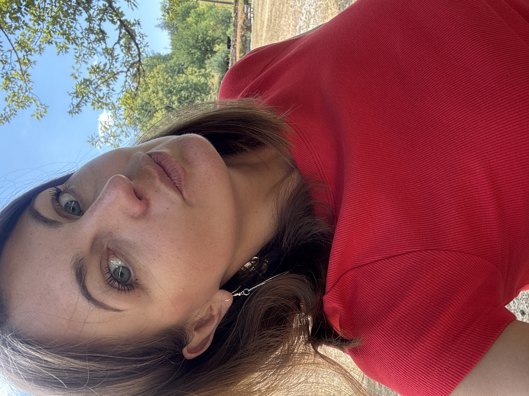 A woman is taking a selfie outdoors in Coteaux du Lizon, France. She has brown hair and is wearing a red top and a silver earring. She is looking at the camera and has her head tilted to the side. The background is blurred, but appears to be a park with trees and a blue sky.