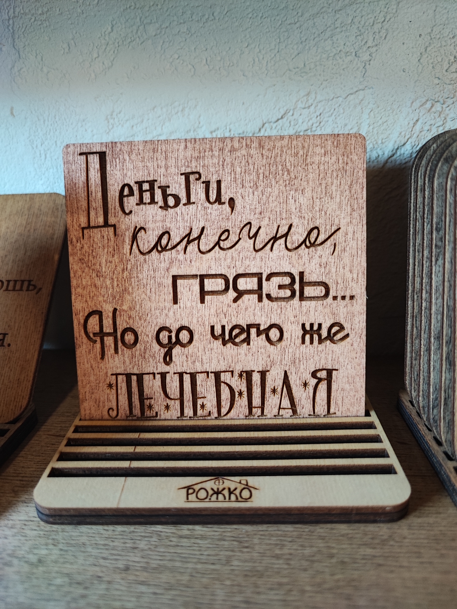 The image shows a wooden plaque with laser-engraved Cyrillic text. The plaque is standing upright on a wooden base with horizontal slats. The text on the plaque reads: "Деньги, конечно, грязь... Но до чего же лечебная". This translates to "Money, of course, is dirt... But how healing it is". Below the plaque, on the wooden base, is a logo that appears to be "РОЖКО". The plaque is positioned on a wooden shelf, and other wooden objects are visible to the left and right of the main plaque, suggesting a display or collection of such items. The background is a textured wall, possibly painted. The lighting is soft, casting subtle shadows. There are no people or animals in the image. The context of the image, specifically its location in Pyatigorsk, Russia, is not visually evident from the image itself.