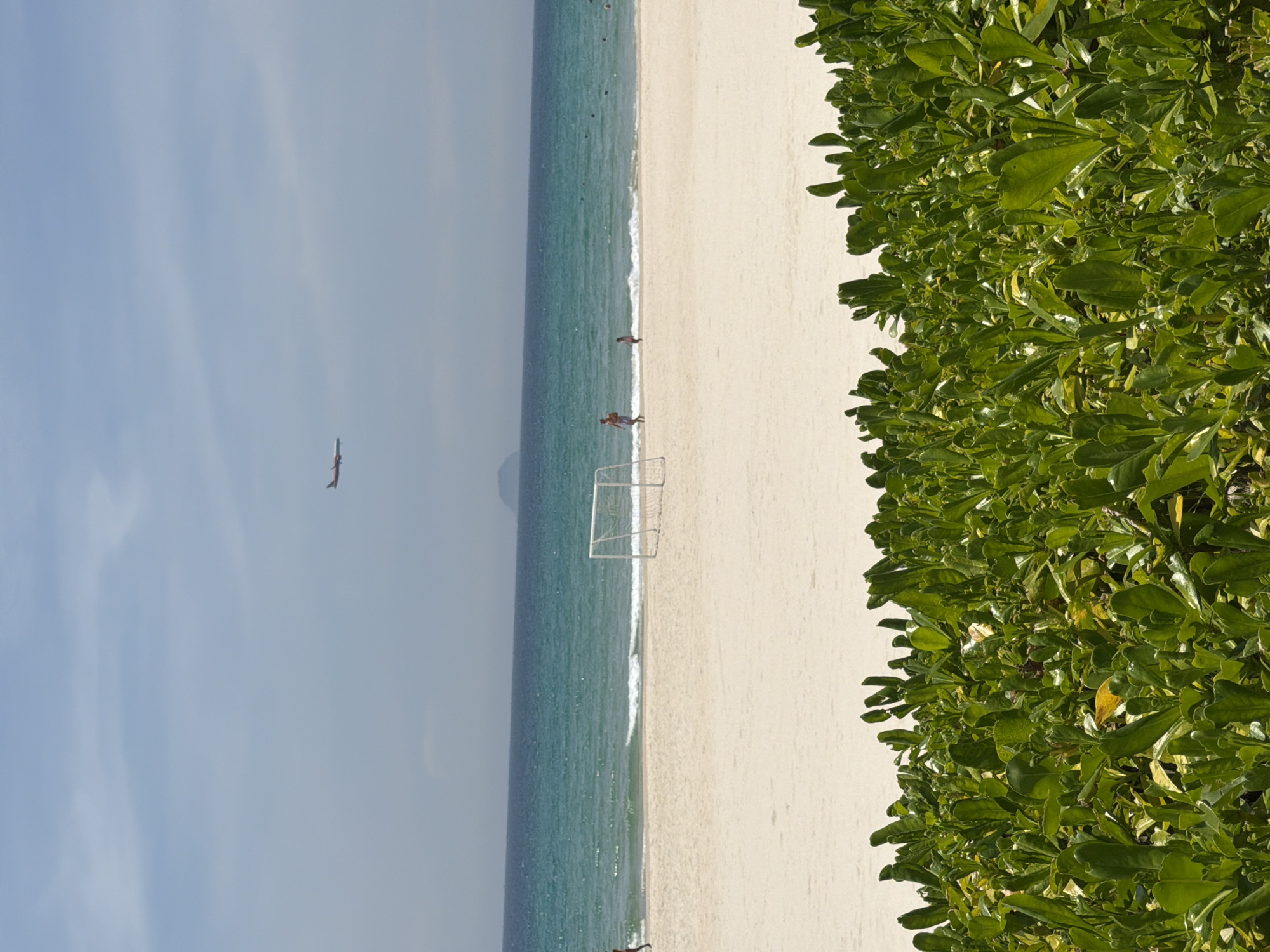 A wide shot captures a beach scene in Cam Lam, Vietnam. In the foreground, lush green foliage dominates the lower portion of the frame. Beyond the foliage, a sandy beach stretches across the mid-ground, with a white soccer goal situated near the waterline. The ocean, a vibrant turquoise and blue, extends to the horizon, where a hazy mountain range is visible in the distance. The sky above is a pale blue with scattered clouds. In the upper part of the sky, a small airplane is in flight. Several people are visible on the beach and in the shallow water, with one person running near the soccer goal. The overall impression is a serene, daytime beach setting.
