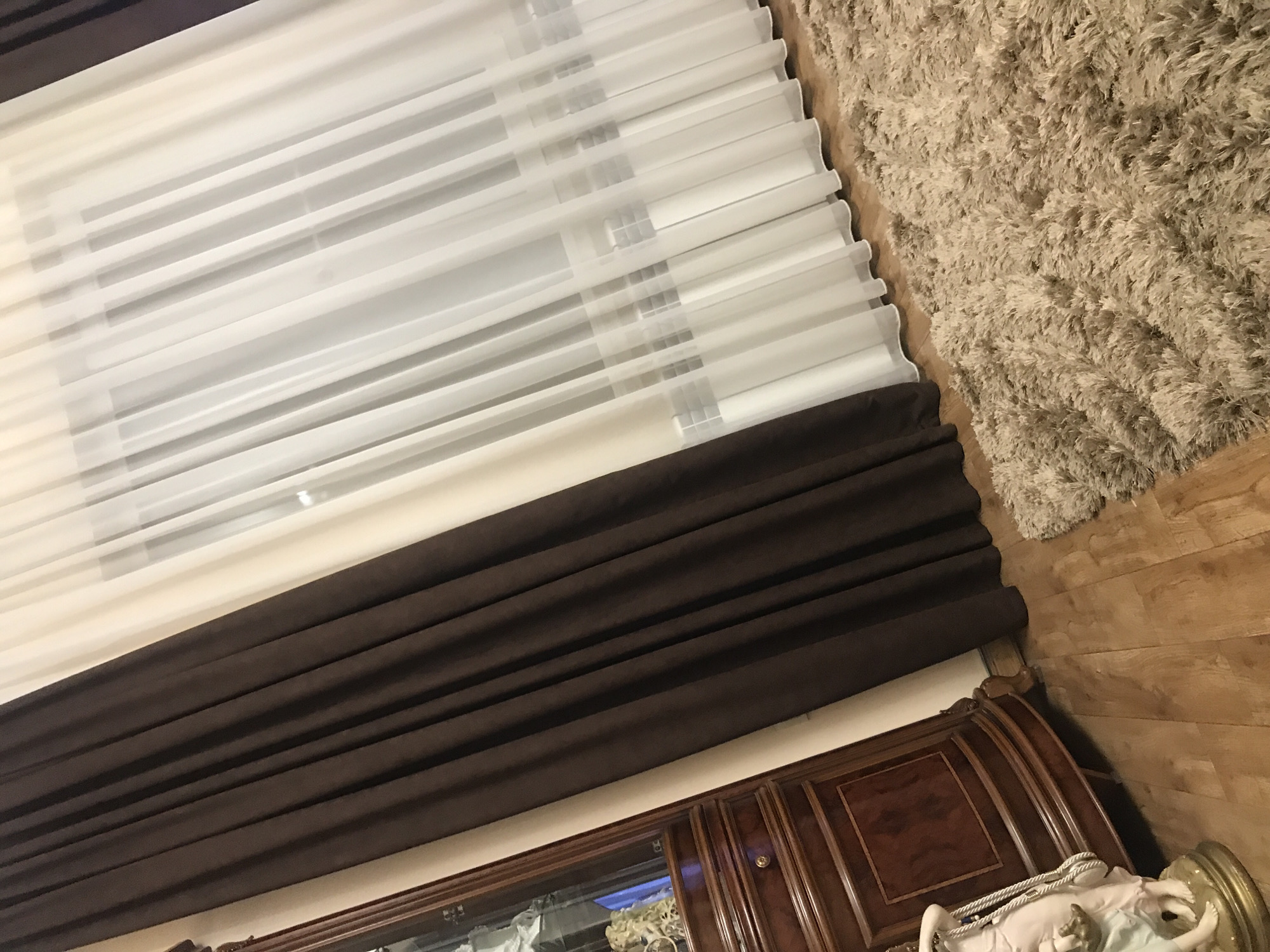 The image displays a partial interior view. At the top left, a window covered with layered, white and translucent blinds. Below the blinds, there's a dark brown curtain, folded in thick pleats.
To the right, a shaggy rug rests partly on the floor. The floor appears to be made of wooden planks.
At the bottom, there's a dark wooden cabinet with glass inlays and ornate detailing. A white statue, mounted on a golden base, is partially visible on the right, placed on top of the cabinet.
There are no visible people or actions within the frame. The location is not discernible from the provided details, and there's no explicit information to confirm it being in Yerevan, Armenia.