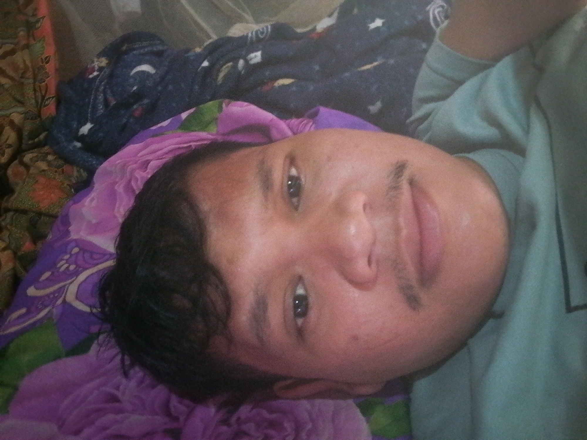 A close-up, low-angle shot shows the face of a young male lying down, his head tilted towards the camera. He has short, dark hair and appears to be wearing a light teal-colored shirt. His left eye is visible, and his mouth is slightly open, revealing his teeth. Behind him, a patterned blanket with blue and purple designs is visible, along with a pink floral-patterned fabric. The scene appears to be indoors, possibly in a bedroom, and the overall lighting suggests it may be day time, but without clear indicators of the time of day. There are no clear landmarks or contextual cues to identify the specific location in Labuhan Bilik, Indonesia.