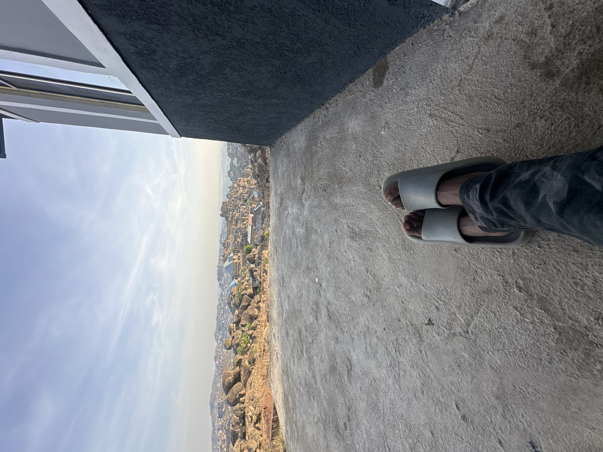 A view of Jos, Nigeria is seen from a high vantage point. The ground nearest to the camera is a textured, concrete surface. A person's feet, wearing light-colored slide sandals, are visible. To the left, there is a structure with a gray textured wall. The distant landscape shows a dense urban area interspersed with rock formations. The sky is lightly clouded.