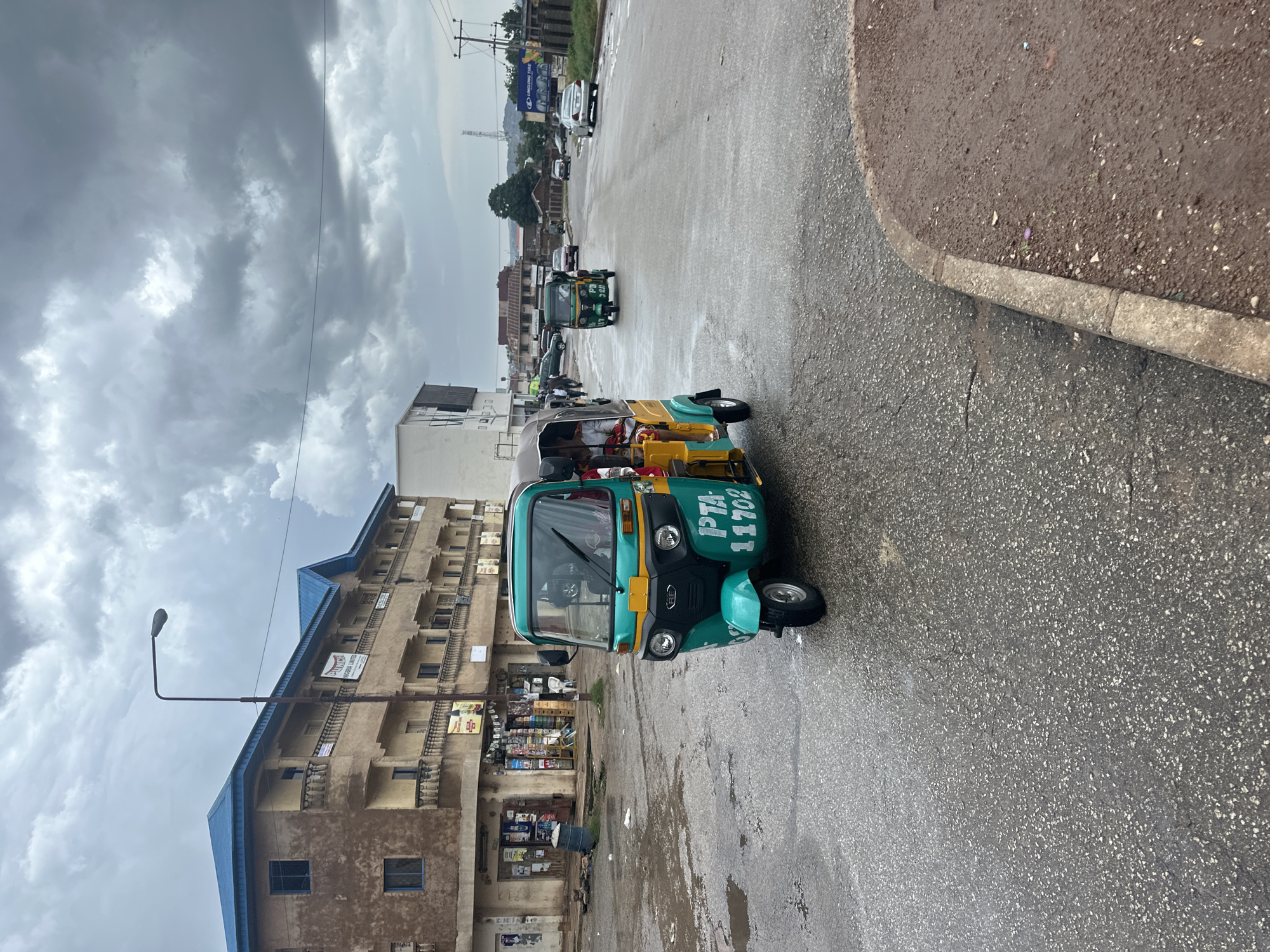 The image captures a street scene in Jos, Nigeria, under a cloudy and overcast sky, suggesting it's either early morning or late afternoon, with the weather appearing damp, possibly after rain. The primary focus is on a row of auto-rickshaws, commonly known as Keke Napeps, parked or driving on a wet asphalt road. One Keke Napep in the foreground is green and yellow, with white lettering on its side that reads "PTA 11702". Other Keke Napeps are visible further down the road, some in motion.
Beside the road, a multi-story building with a distinctive blue roof stands out. The building has a weathered, beige facade and balconies, and appears to house shops or businesses on its ground floor, evidenced by visible merchandise and displays. A street lamp on a tall pole extends from the left side of the image towards the building. In the background, more buildings and utility poles are visible, hinting at a moderately developed urban environment. The overall atmosphere is one of daily life and commerce continuing despite the dull weather. There are no visible people clearly identifiable in the image, but their presence is implied by the vehicles and the active street.