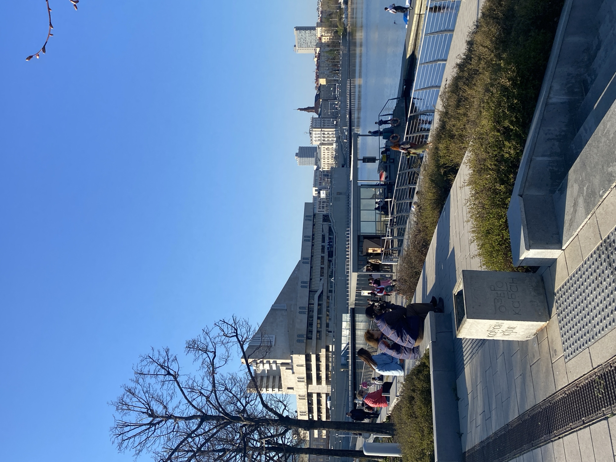 The image shows a waterfront scene in Kazan, Russia. The background features a river with city buildings visible on the opposite bank. In the foreground, there is a paved walkway with people sitting along a low wall and on benches. A square concrete block is visible on the walkway. A bare tree is on the left side of the frame, against a clear blue sky. The focus is sharp and the lighting is bright.
