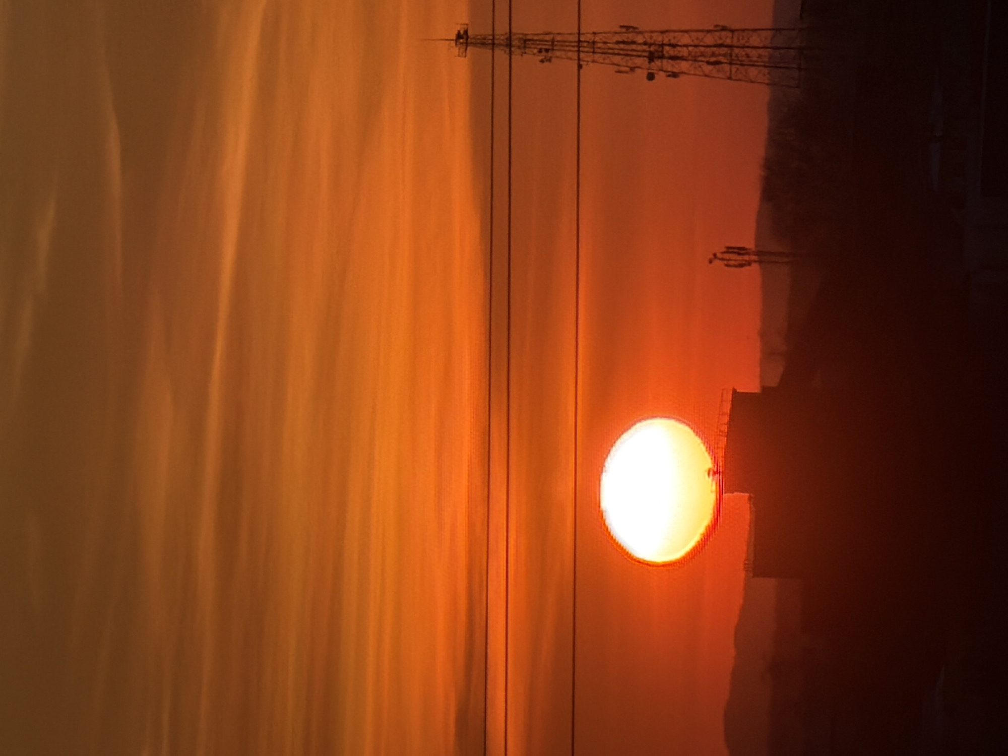 The image depicts a sunset in an urban environment. The sun is prominently positioned, appearing as a bright orb partially obscured by a dark building or structure on the right side of the frame. Silhouetted against the sky, a communication tower stands to the left of the sun. The sky displays a warm orange hue, suggesting either sunrise or sunset conditions. In the city of Chaikovsky, Russia, the environment includes utility poles.