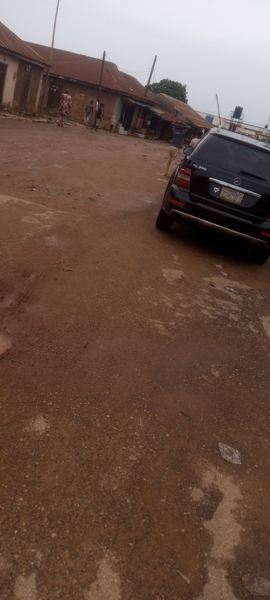 The image shows a dirt road in Sabon Tasha, Nigeria. A black Mercedes-Benz ML 350 is parked on the right side of the road, partially blocking the view. Several people are standing near the buildings across the road. The buildings appear to be simple, single-story structures with rusted roofs. The environment seems muddy, suggesting recent rainfall.