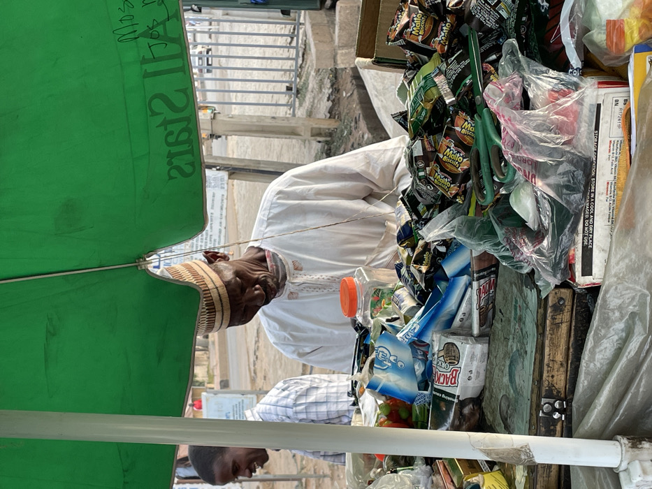 An elderly man wearing a traditional cap and white robe stands behind a stall laden with various packaged goods, including "Mallo" biscuits and other snacks. The stall is covered by a green umbrella. The writing "ALL STARS MOS" is visible on the umbrella. A younger man in a checkered shirt is partially visible in the foreground. Some text, including "STORAGE CONDITION: STORE IN A COOL AND DRY PLACE" and "IF DAMAGED, DO NOT USE," can be seen on one of the boxes.