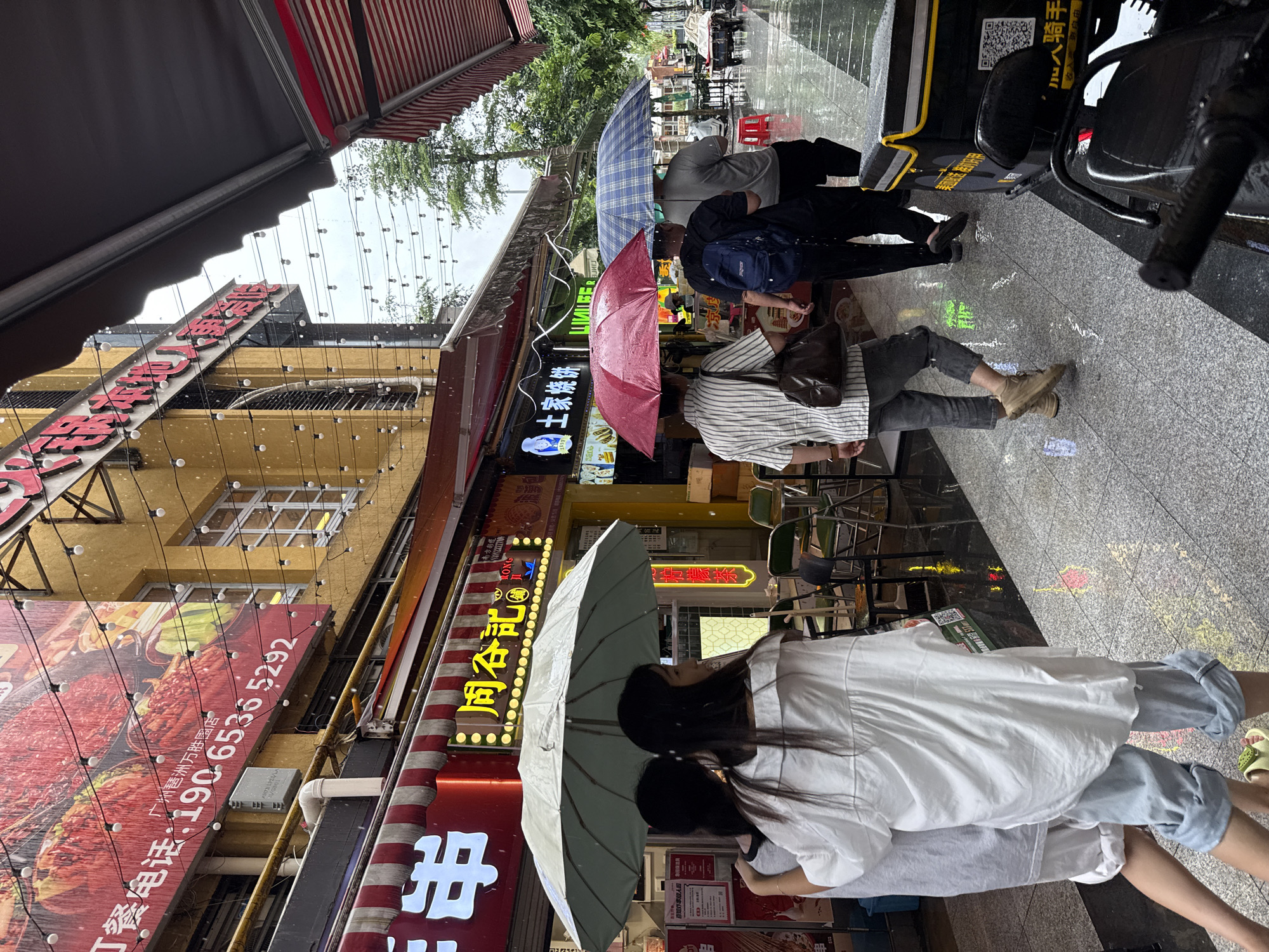 The image depicts a street scene in Haizhu District, China, during a rain shower. Several people are walking under umbrellas, while others are walking without protection from the rain. Shops with awnings line the street, some displaying large advertisements. The ground is wet and reflects the lights from the shops and string lights overhead. A delivery scooter is parked on the right side of the image. The overall atmosphere is that of a bustling street encountering inclement weather.