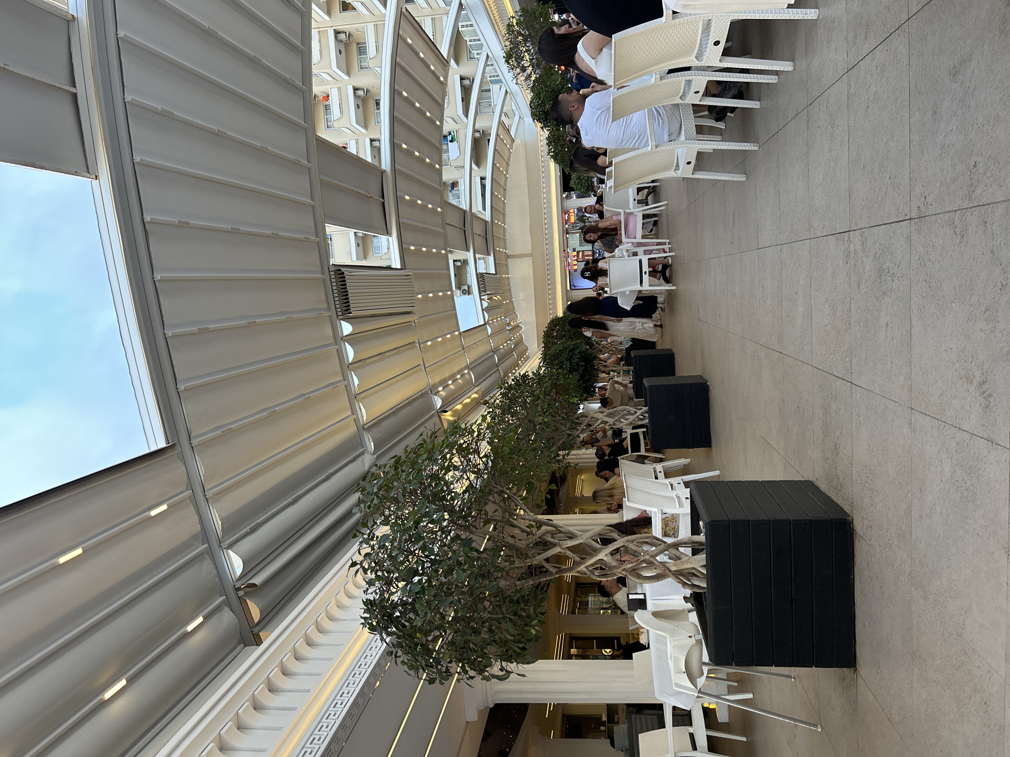 The image depicts an outdoor restaurant or cafe setting. Patrons are seated at tables with white chairs. Trees in black planters line the dining area. An overhead retractable awning covers the space. The location appears to be in Unknown, Unknown. The scene is well-lit.