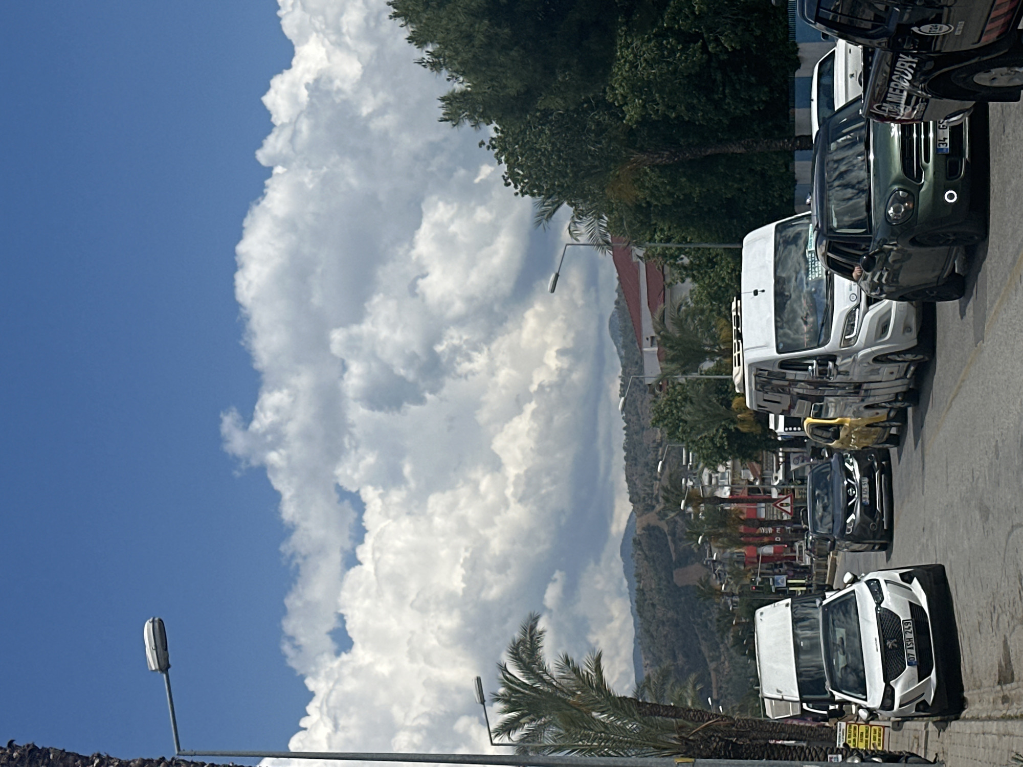 The image shows a street scene in Fethiye, Turkey, characterized by a combination of natural and urban elements.
In the foreground, several vehicles are parked along the right side of the road. They include vans, a dark green Mini Cooper, and a black SUV. The vehicles face towards the top of the frame.
On the left, a blue sky dominates, filled with cumulus clouds.
In the background, a mountain range is visible, partially obscured by buildings, palm trees, and trees. Streetlights are situated along the road.