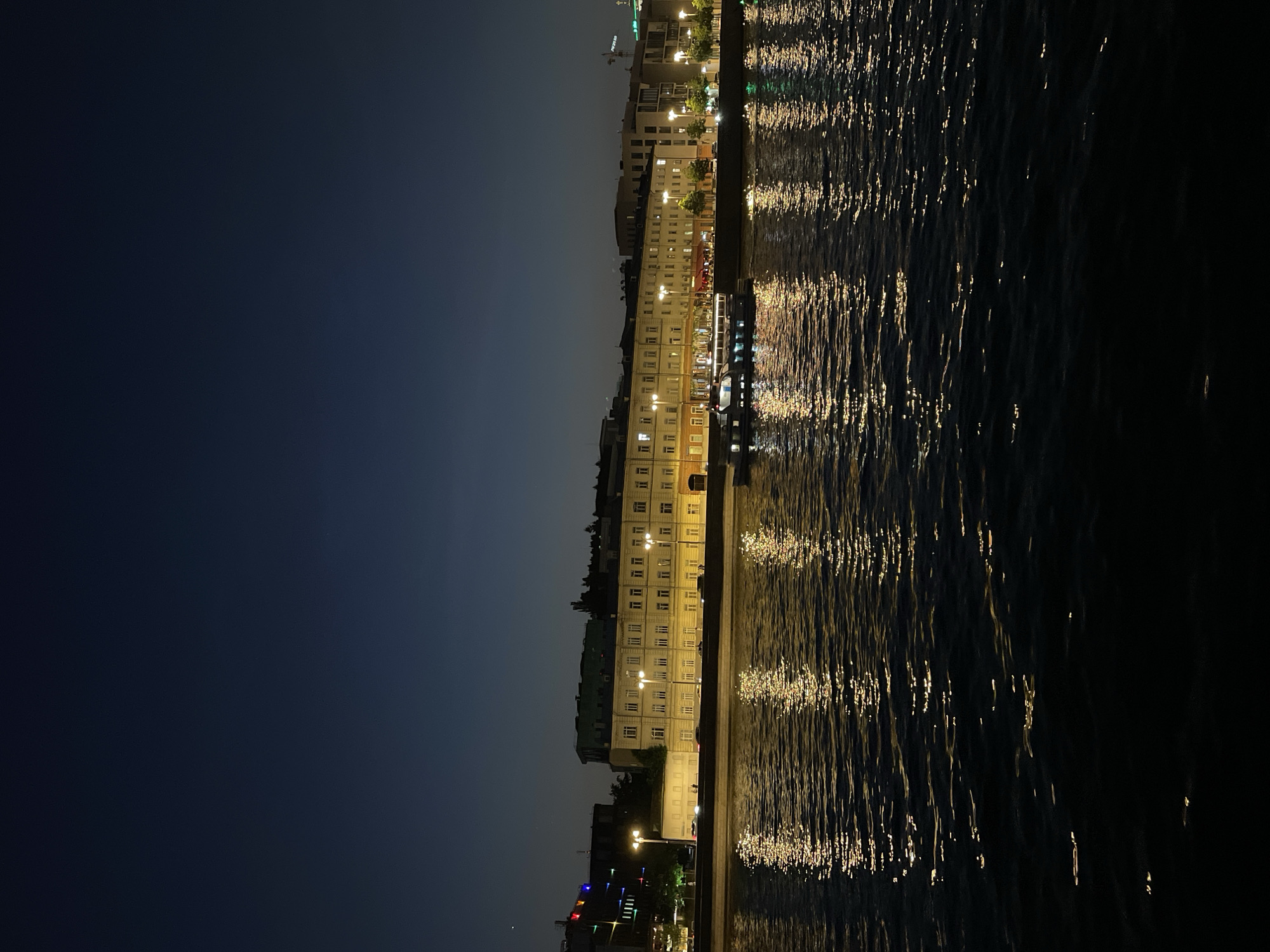 The image shows a nighttime view of a cityscape along a body of water, likely in Moscow, Russia, based on the architectural style of the buildings. The lighting is primarily from street lamps and building illumination, reflecting in the water's surface. The sky is dark with a gradient towards the horizon. The buildings appear to be old and have many windows. There are no people visible. The water is dark with light shimmering across its surface. The photograph is taken from a low angle and is rotated to make it vertical.