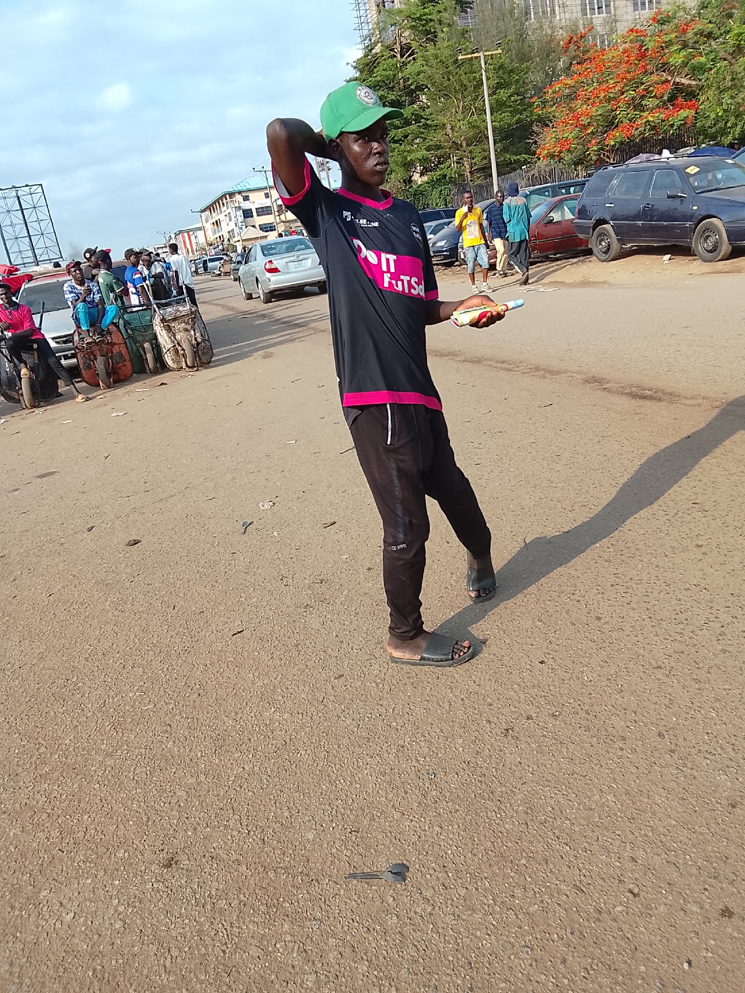 A man is standing on a street in what appears to be a city, likely Abuja, Nigeria, based on the style of vehicles and structures. He is wearing a black t-shirt with pink accents, black pants, sandals, and a green baseball cap. He is holding a roll of what looks like money or tickets in his left hand and has his right hand on the back of his head. In the background, there are cars, pedestrians, trees, and buildings. Some individuals are gathered around handcarts. The road surface is rough asphalt, and a small object resembling a key lies on the ground nearby. The lighting suggests it is daytime, with a long shadow cast by the standing man.