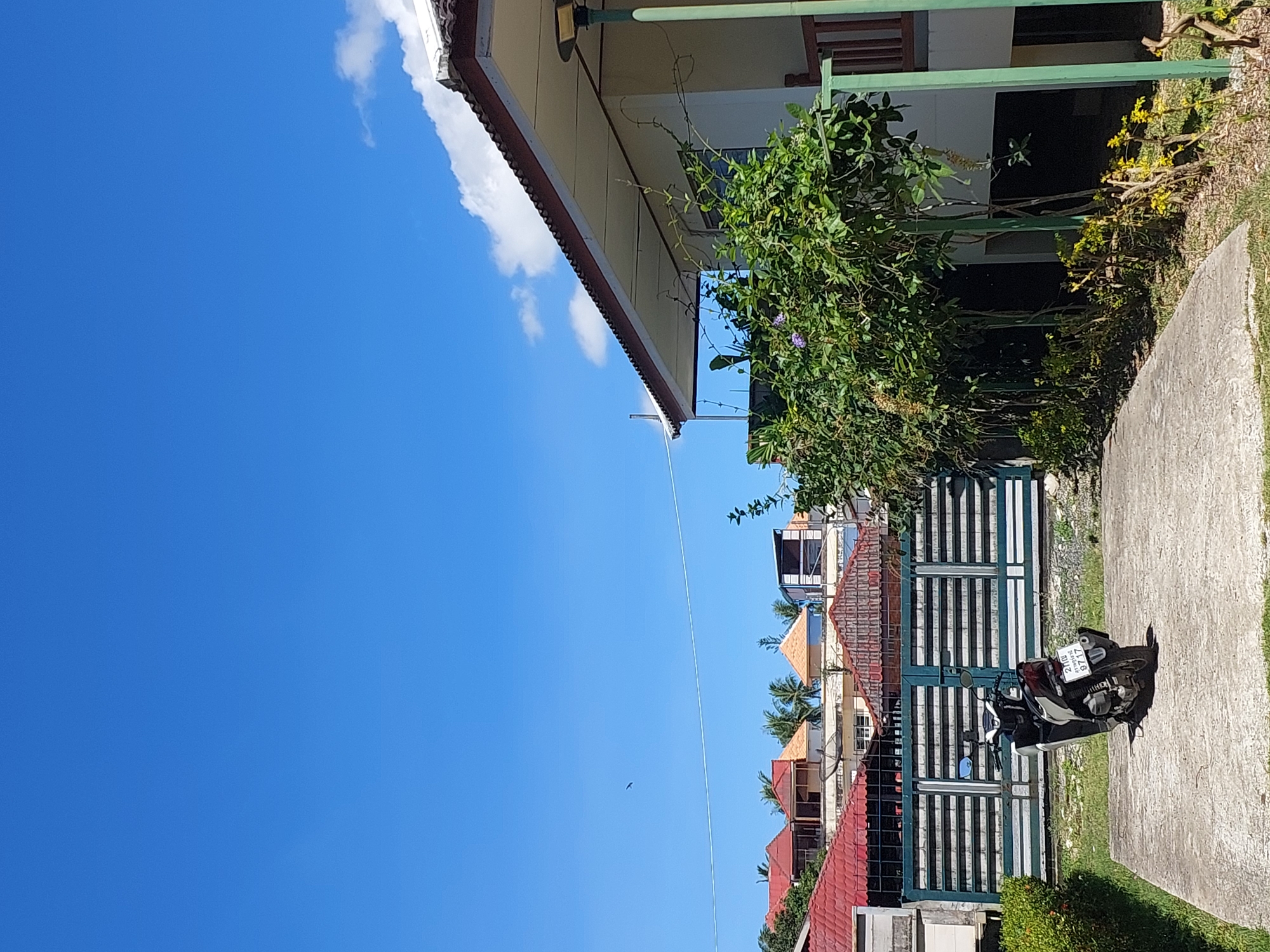 The image depicts a scene in Baan Mae Nam, Thailand, under a clear blue sky with scattered white clouds. In the foreground, a concrete pad is visible, with a silver and black scooter parked on it, facing away from the camera. The scooter has a license plate with Thai characters and the number "8717". Behind the scooter is a large, green metal gate with horizontal bars and decorative elements. Beyond the gate, several buildings with red and orange tiled roofs are visible, along with palm trees. To the right of the scene, a building with a white exterior and a light brown roof with dark brown trim is partially visible. A lush green bush with some small purple flowers is in front of this building, and a tall green pole with a light fixture is to its right. The overall environment suggests a residential or semi-commercial area in a tropical location.