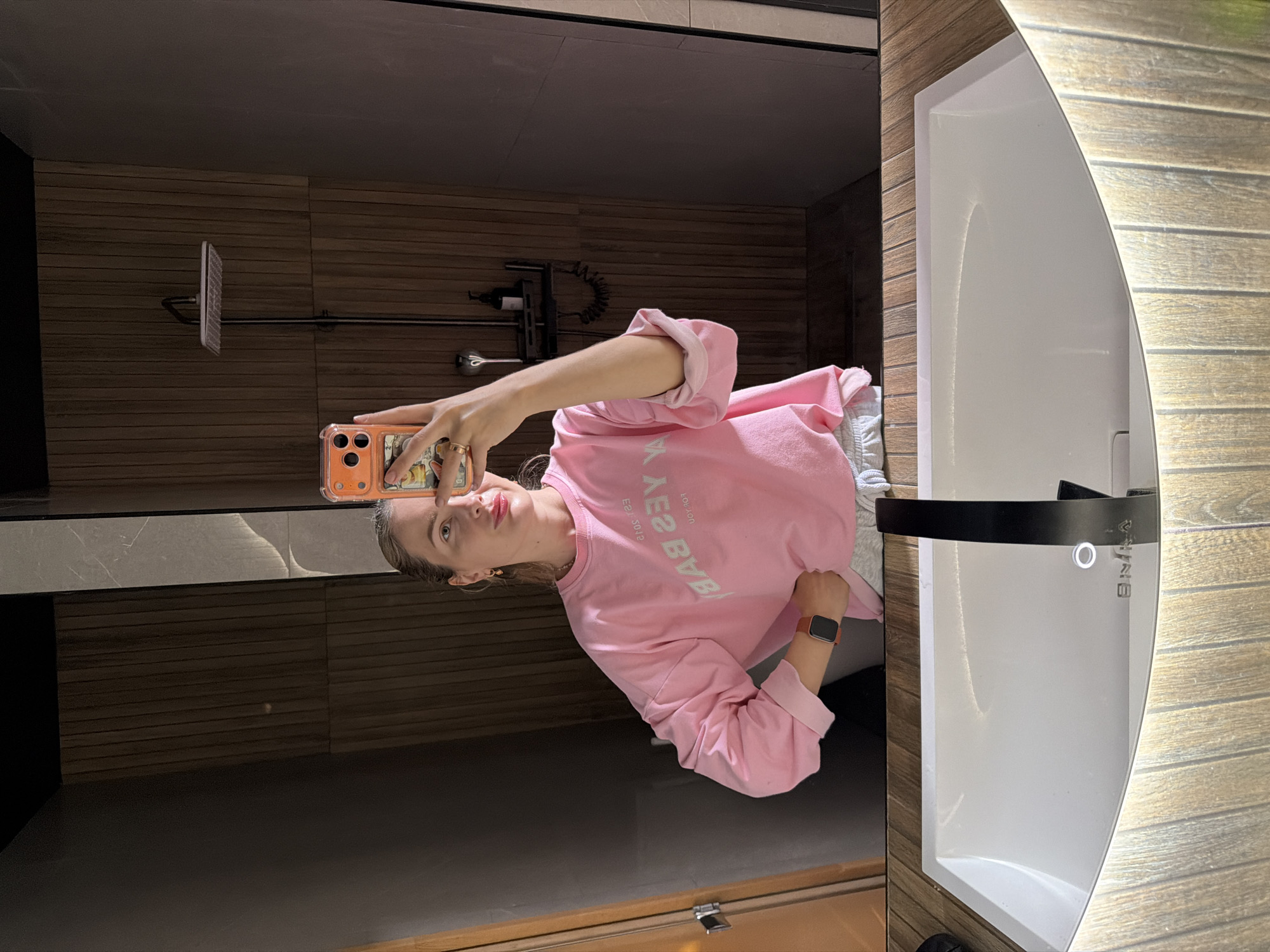 A young woman is taking a mirror selfie in a bathroom. She is wearing a pink sweatshirt and light-colored shorts. Her hair is pulled back in a ponytail. She has on makeup, including red lipstick. She is wearing a watch on her left wrist and a ring on her right hand. The bathroom has wooden walls and a modern sink with a black faucet. The scene is located in a bathroom in the city of Semiluki, Russia.