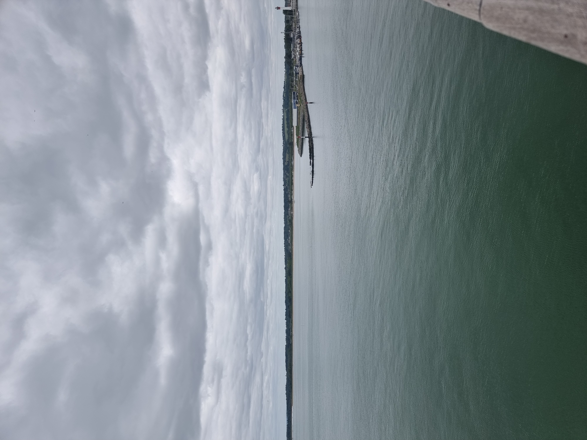 The image captures an expansive, elevated view of the harbor entrance and bay in Ouistreham, France, under a heavily overcast sky. The upper two-thirds of the frame are dominated by a dense canopy of textured grey clouds, with no clear sky visible. Small, dark specks are sparsely distributed across the sky. The lower portion of the image consists of a calm, greenish-grey body of water, displaying subtle ripples across its surface.
A distinct horizon separates the water from a distant, low-lying landmass. This landmass stretches across the entire width of the mid-ground, characterized by a continuous dark green band of trees and vegetation along its edge, with fainter indications of land beyond.
On the right side of the image, a prominent man-made structure, a dark, rocky breakwater or jetty, extends into the water. At the far end of this breakwater, a small, red and white striped lighthouse or navigation beacon stands. Another similar, smaller beacon is positioned further back along the breakwater. Behind the breakwater, a marina is visible, densely populated with numerous small to medium-sized boats docked in their slips, flanked by light-colored harbor buildings along the shore. Further to the right, a tall, distinctive red and white striped lighthouse serves as a prominent landmark for the harbor entrance.
In the bottom right corner, a portion of a weathered, grey wooden structure, likely a railing or deck edge, is visible, indicating the photograph was taken from an elevated vantage point, possibly from a ship or pier. There are no discernible people or active interactions visible in the scene.