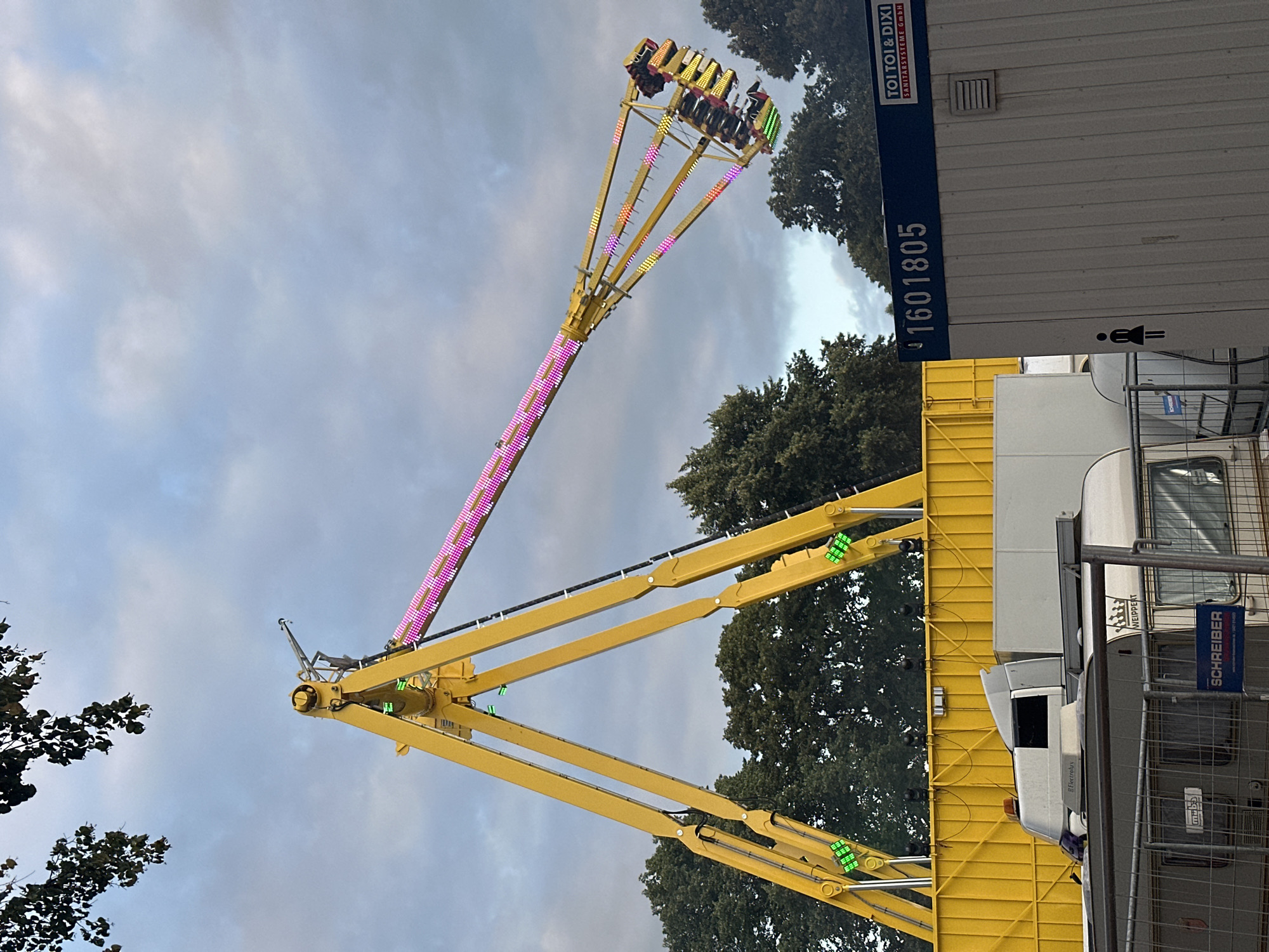 A large yellow amusement park ride, likely a pendulum ride, is illuminated with pink and yellow lights and is in motion against a cloudy sky. The ride has a central A-frame structure supporting a long arm that swings passengers in a gondola. Several people are visible in the gondola. In the foreground, there are several portable toilet units, one of which has the branding "TOI TOI & DIXI SANITÄRSYSTEME GMBH" and the number "1601805" printed on it. There are also some caravans and a temporary fence. Trees can be seen in the background. The image was taken in Bruchhausen-Vilsen, Germany.