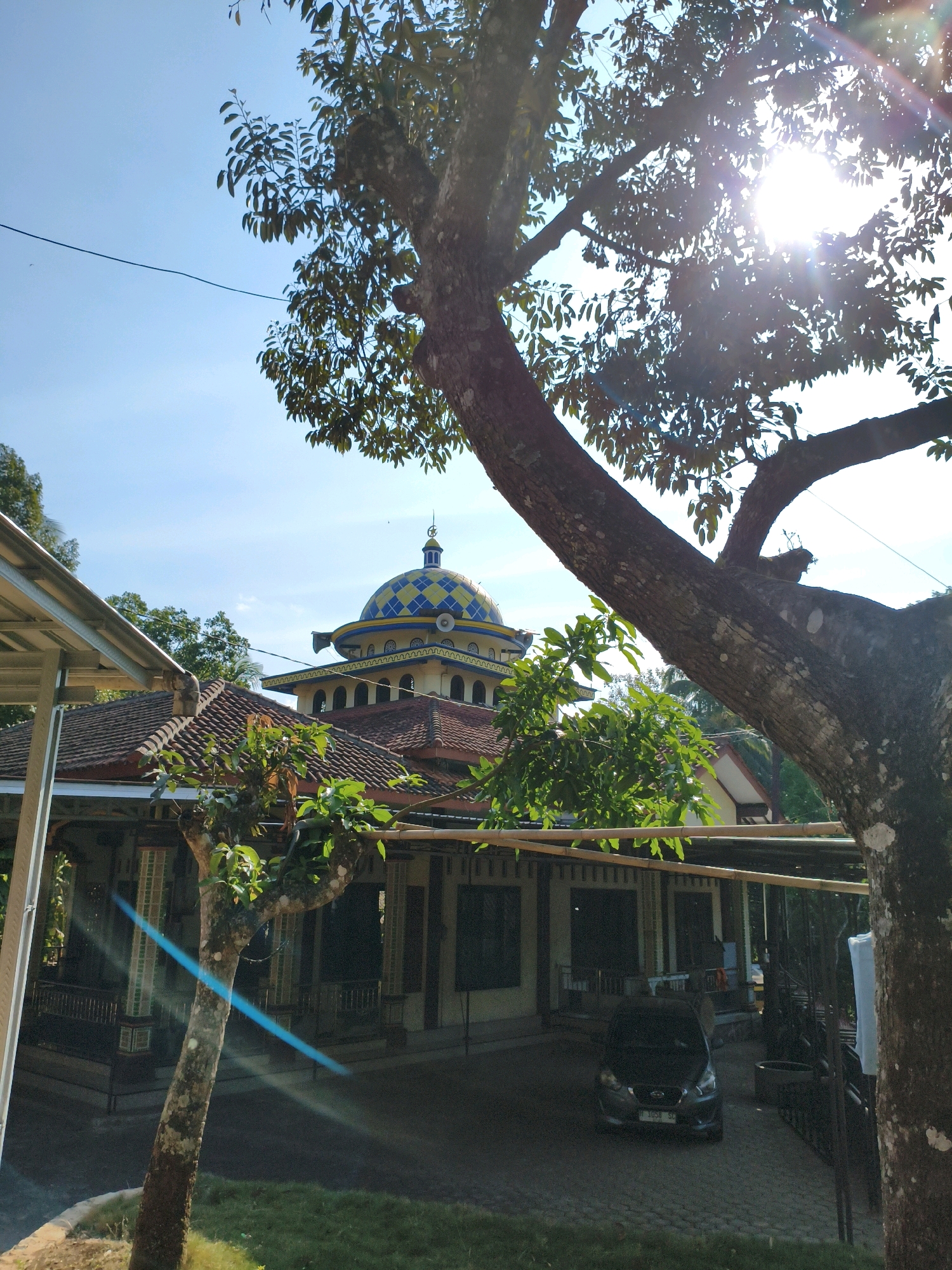 The image shows a mosque in Segiri, Indonesia, with a distinctive blue and yellow checkered dome. The mosque appears to be a single-story building with a tiled roof, partially obscured by trees in the foreground. A dark-colored car is parked in front of the mosque. The sun is shining brightly, casting lens flares across the scene. The sky is clear blue. The overall impression is one of a sunny day in a tropical environment.