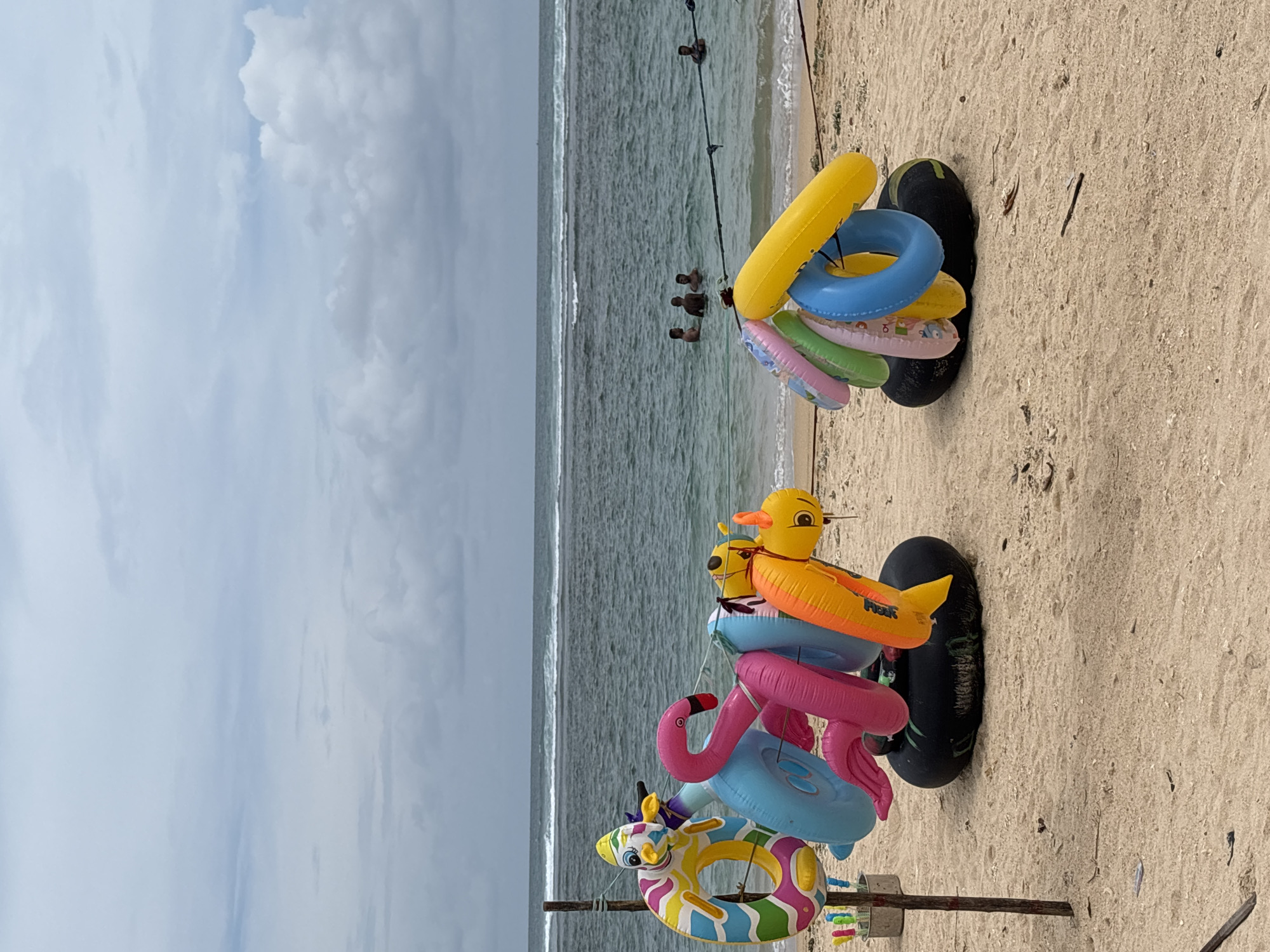 The image shows a beach scene in Weliweriya (SP), Sri Lanka. Inflatable swimming aids, including rings and animal shapes (duck, flamingo, zebra), are stacked on black inner tubes standing on the sand. Several people are visible in the ocean's shallow water. The sky is cloudy.