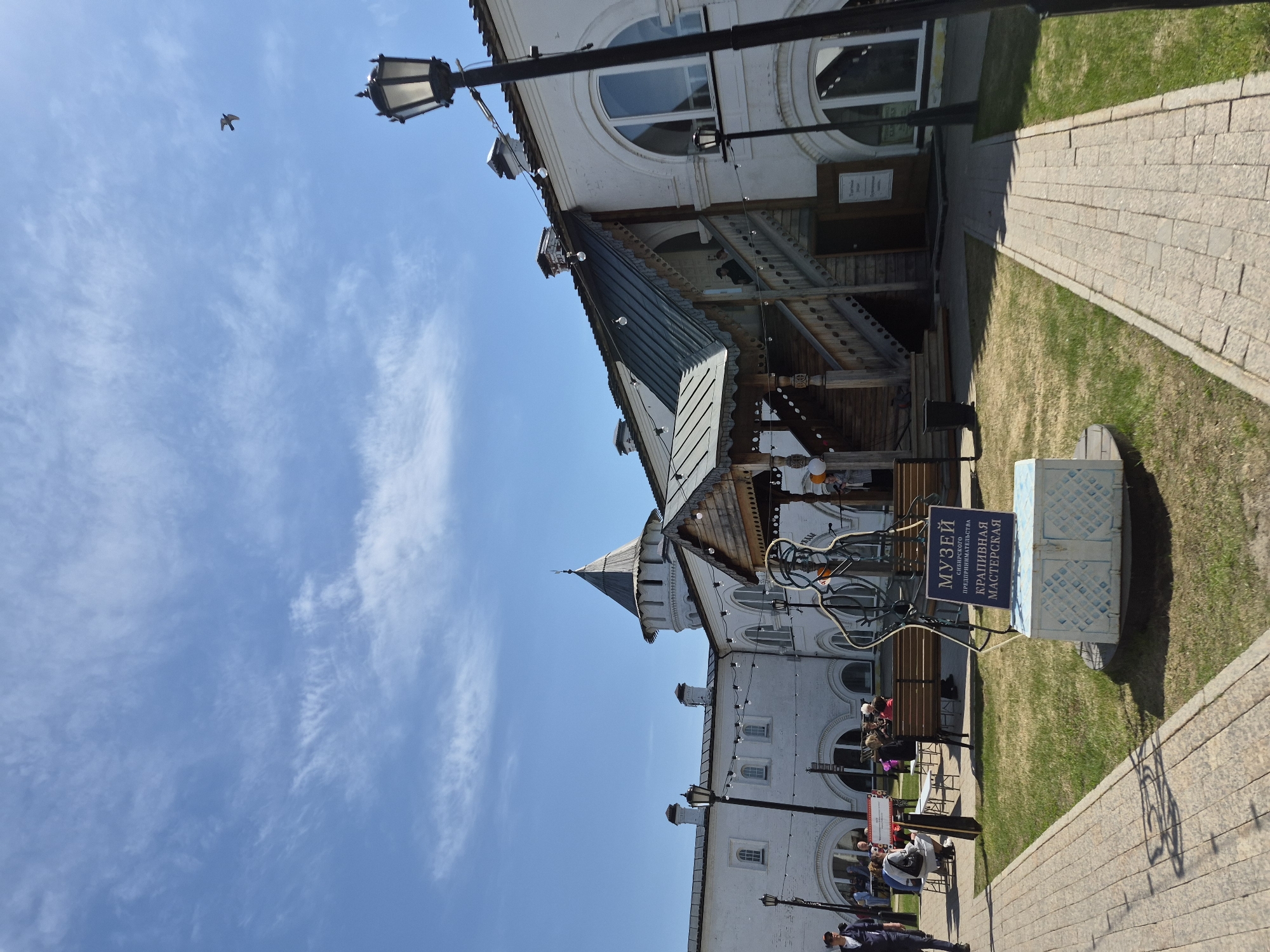 The image shows a street scene in Tobolsk, Russia, featuring the Museum of Siberian Entrepreneurship Craft Workshop (МУЗЕЙ ПРЕДПРИНИМАТЕЛЬСТВА СИБИРСКОГО КРАПИВНАЯ МАСТЕРСКАЯ). The foreground contains a stone-paved walkway, a patch of grass, and a sign indicating the location of the museum. People are seated at tables under the building, while others are walking by. The architecture is primarily white with arched windows and a complex roof structure. A bird is seen in flight in the blue sky. A black lamppost is visible in the frame.