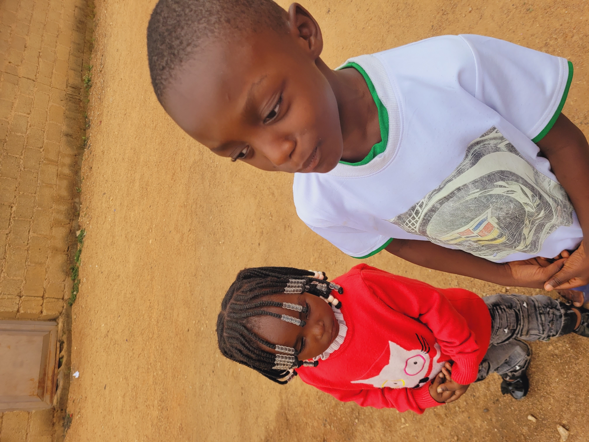 Two young children are the main subjects of the image, positioned outdoors on a dirt ground near a brick wall. The older child, a boy, is wearing a white t-shirt with green trim and a graphic design. He is looking down at the younger child. The younger child, a girl, is wearing a red sweater with a white cat graphic. Her hair is styled in braids, adorned with beads. They appear to be standing together. The location is presumed to be Bukuru, Nigeria.