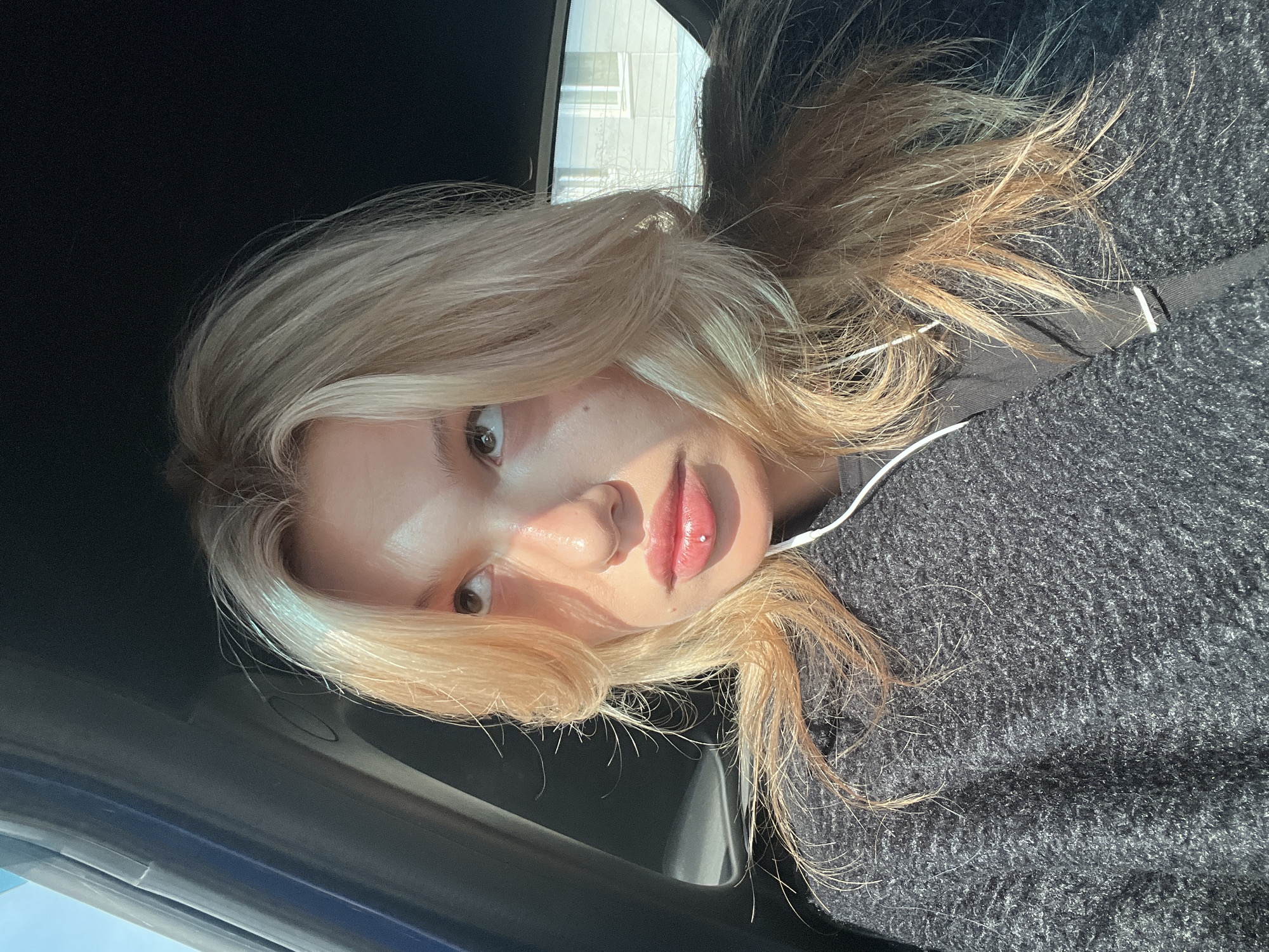 A close-up, angled portrait of a young, fair-skinned individual with blonde hair. The person has light-colored eyes, and a lip piercing is visible on their lower lip. They are wearing a dark gray or black sweater. The image appears to be taken inside a vehicle, based on the presence of the car's interior visible in the top left and lower left corners of the frame. Light streams in from an outside source, creating bright highlights and shadows on the person's face. Out the car window, one can see a light-colored building. Due to the building style, it is plausible the photo was taken in Chaikovsky, Russia.
