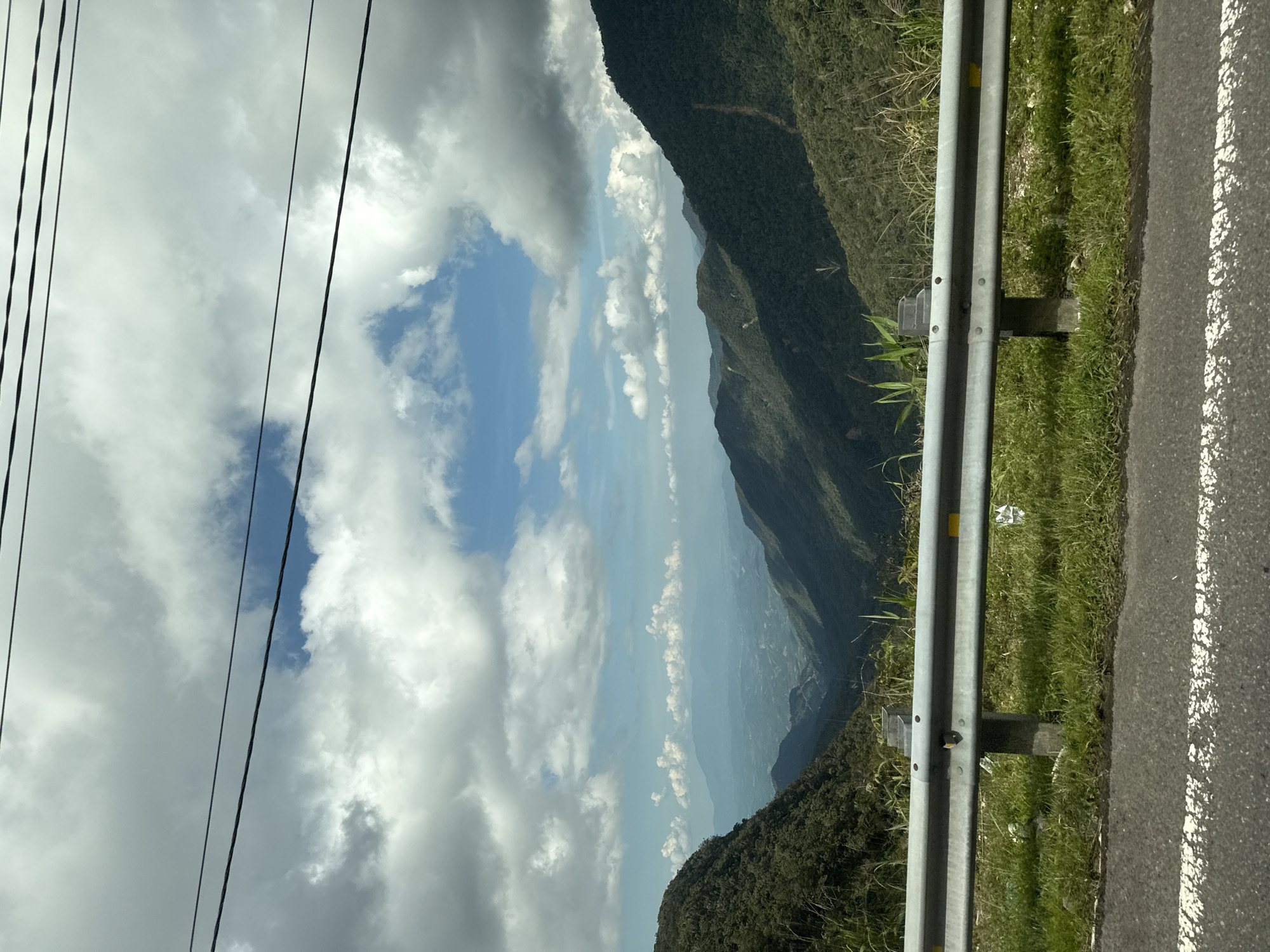 The image depicts a scenic mountain landscape with a valley and distant hills under a cloudy sky. The foreground shows a grey metal guardrail with posts, situated on a grassy verge next to an asphalt road with a white line. Beyond the guardrail, the view opens up to a lush green valley, with successive layers of mountains fading into the distance, bathed in sunlight and shadow. The sky is filled with dramatic, cumulus clouds, with patches of blue sky visible. The scene appears to be a view from a road winding through a mountainous region, possibly in the vicinity of Nam Khánh Vĩnh, Vietnam, given the terrain and vegetation. There are no people or specific objects other than the road infrastructure visible in the scene.