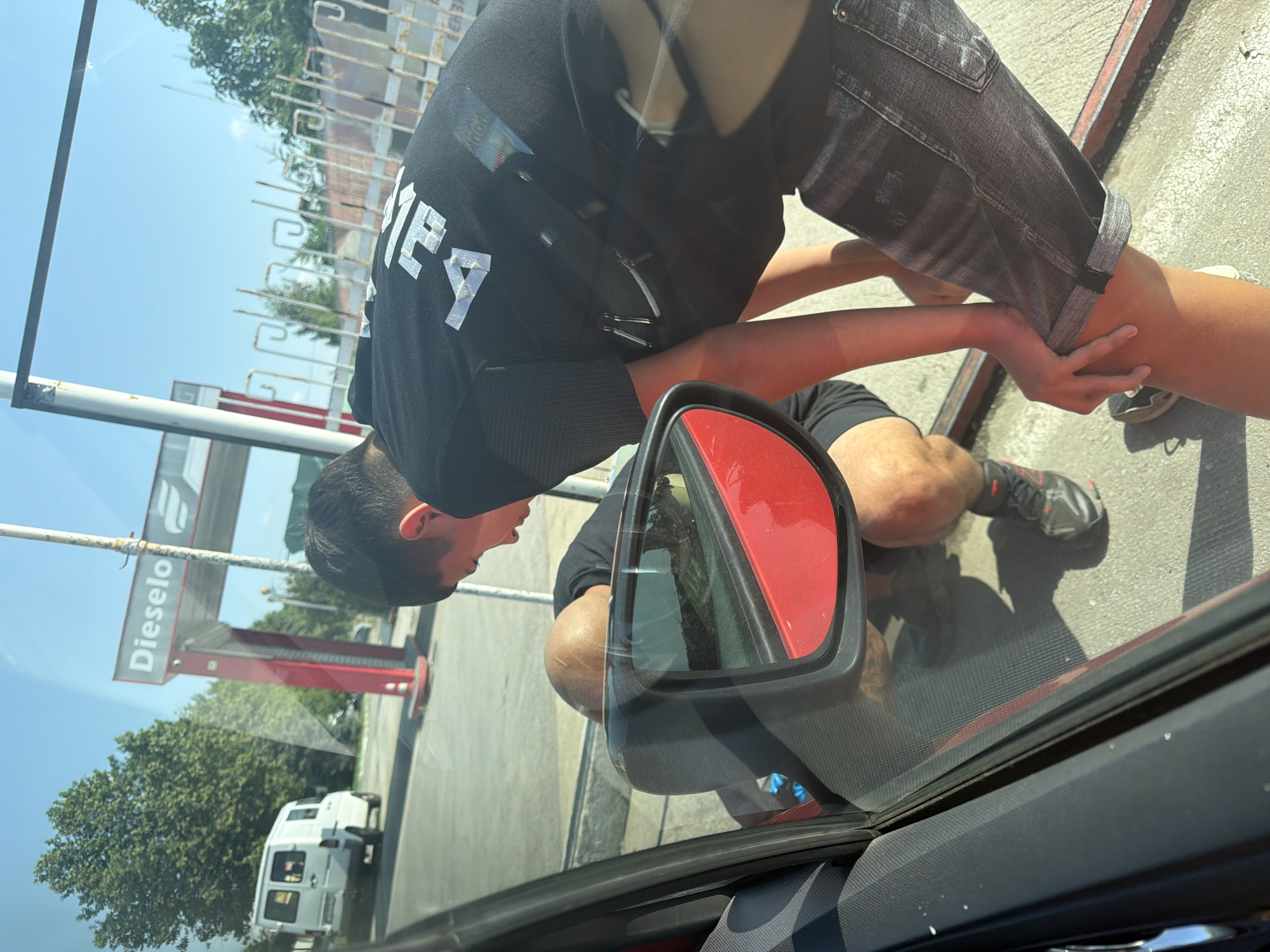 The image, when correctly oriented, captures an outdoor scene at what appears to be a gas station during bright daylight. A person with dark hair, wearing a black t-shirt with fragmented white text ("ME4" or similar) and dark denim shorts, is leaning into the driver's side of a red car. Their posture suggests they are interacting with something or someone inside the vehicle. A second pair of hands, with a lighter skin tone, is visible holding the first person's leg just above the knee, implying another individual is seated inside the car.
Key objects include the red car's side mirror, which reflects parts of the scene, and the car's window and door frame. In the background, a large red and white sign for a fuel pump is visible, clearly displaying the word "Dieselo," indicating a diesel fuel station. A gas pump structure, a white van, and several green trees are also seen. A metal fence runs across the mid-ground.
The time of day is sunny, likely midday, given the clear blue sky and strong shadows cast on the ground. No particular emotions are discernible from the subjects. The setting is clearly a fuel station or similar roadside service area.