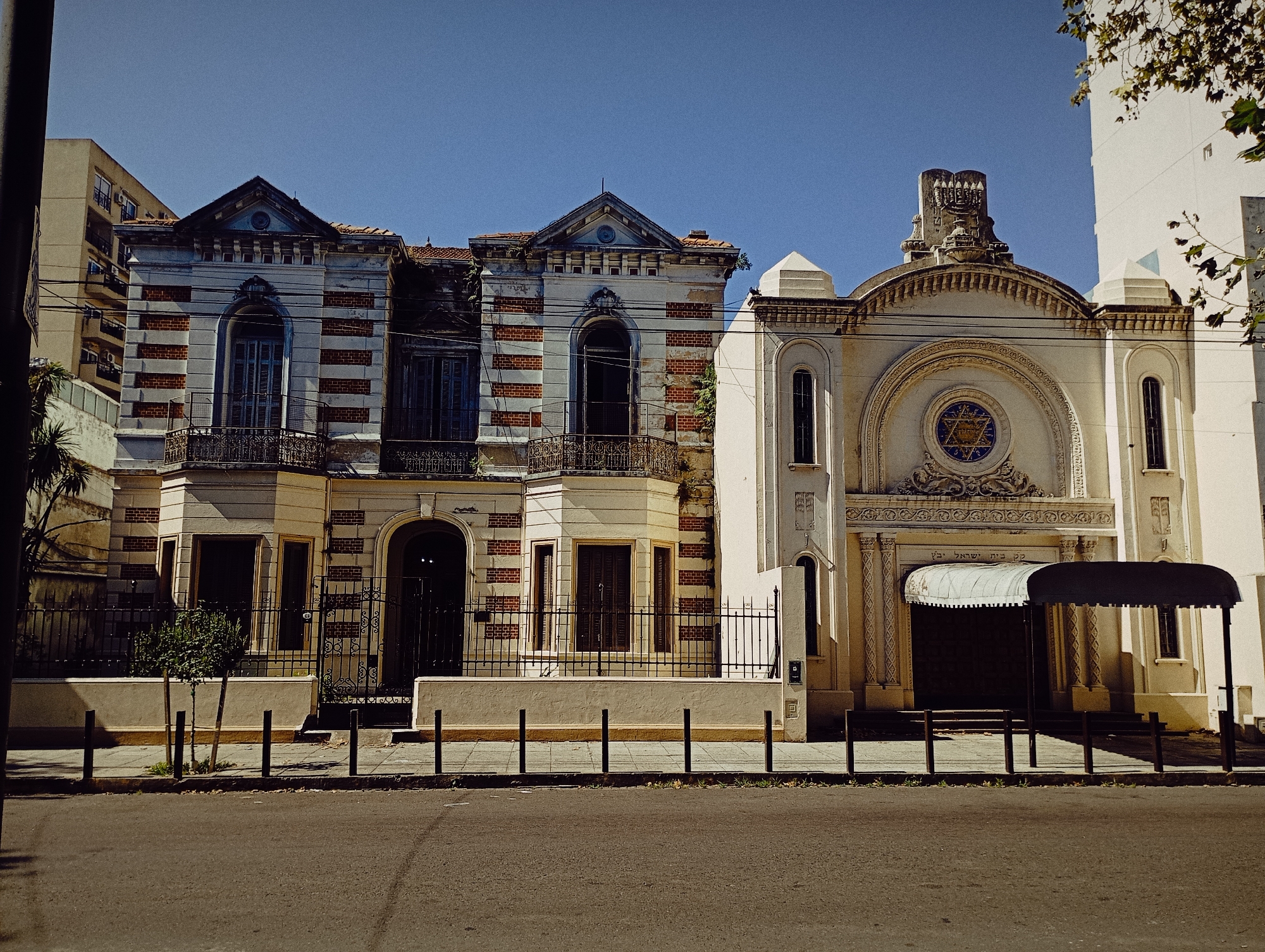 The image depicts a street scene in Buenos Aires, Argentina, featuring two adjacent buildings with distinctive architectural styles. The building on the left displays a deteriorating facade with a blend of yellow and brick-lined sections, ornate balconies, arched windows with closed shutters, and visible signs of decay. To the right stands a synagogue characterized by its cream-colored facade, a large Star of David window, and a covered entrance. The street in front is paved with asphalt and has a series of bollards separating the sidewalk from the road. Overhead, clear blue skies dominate the background. A tall, modern building with exposed air conditioning units is on the left. Parts of a tree are present on the right side. A row of bollards separates the sidewalk from the street.