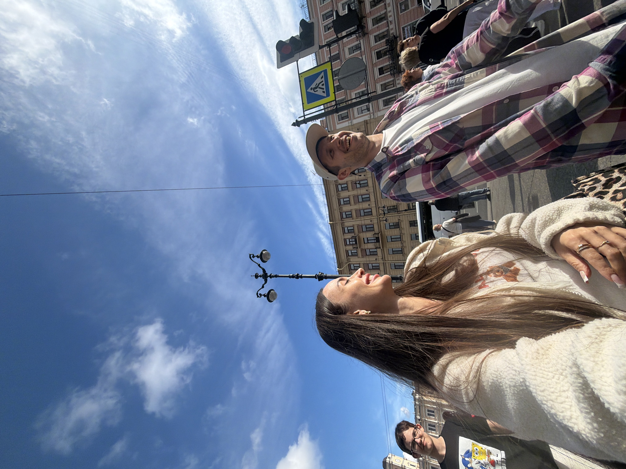 The image shows a group of people in an urban setting with a bright blue sky and scattered clouds. In the foreground, a woman with long brown hair is smiling, wearing a cream-colored fuzzy jacket with a graphic print on it. She is wearing rings on her fingers. Beside her, a man with short brown hair and glasses is looking towards the camera with a slight smile, wearing a black t-shirt with a cartoon print.
Further back and to the left, a man wearing a plaid shirt and a baseball cap is laughing. Behind him, other individuals are visible, some walking and some standing, with buildings lining the street. Traffic lights and a pedestrian crossing sign are present, suggesting an intersection.
The overall scene appears to be outdoors during the daytime, likely a sunny day given the clear sky and bright lighting. The architecture of the buildings suggests a European city, and the provided location context identifies it as Saint Petersburg, Russia. No discernible text is clearly legible in the image, beyond the cartoon print on the t-shirt and potentially on the clothing of others.
