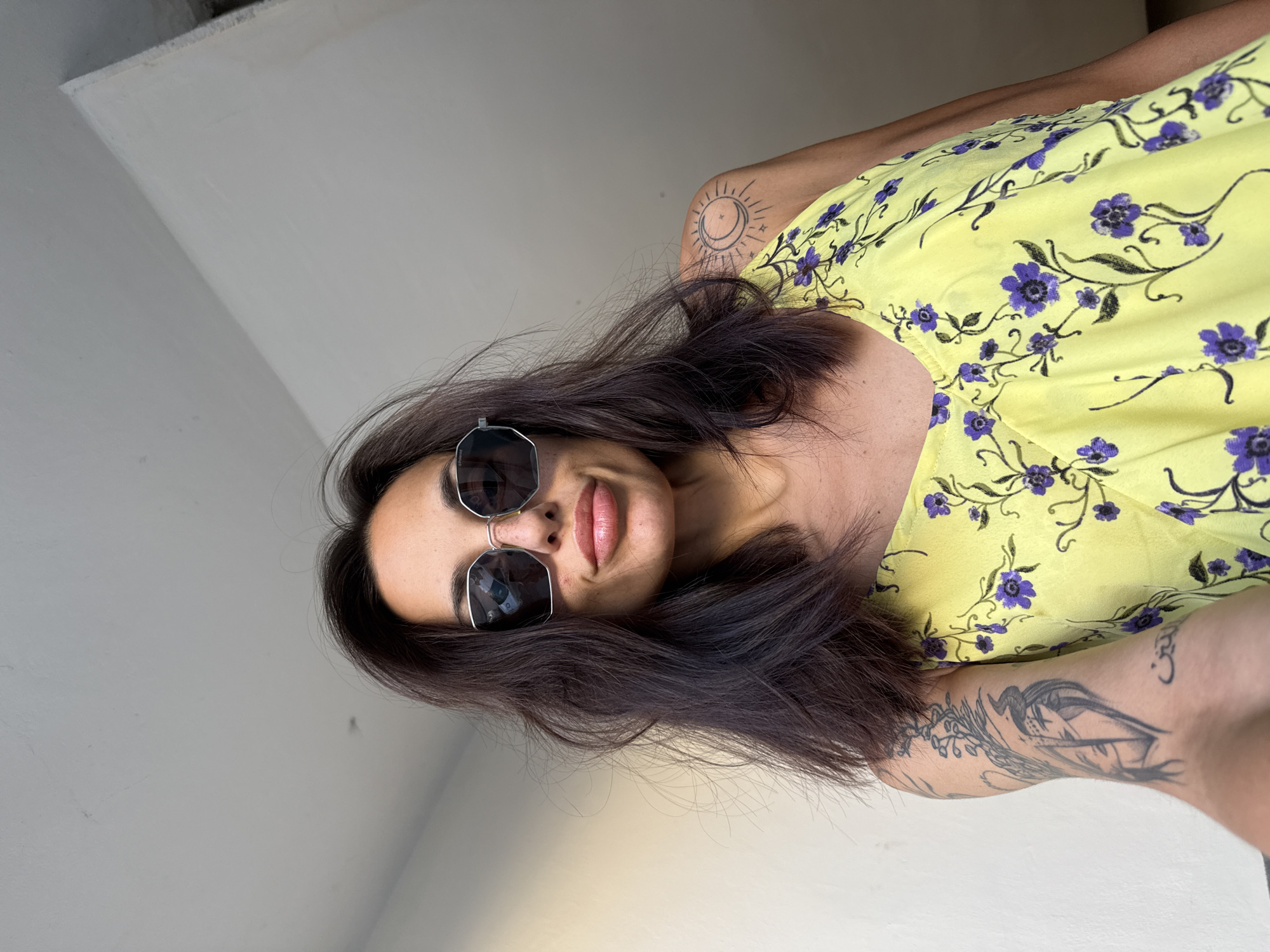 A woman with dark hair and sunglasses poses for a selfie indoors. She is wearing a yellow dress with a floral pattern. She has tattoos on her arm and shoulder. The background is a plain wall and ceiling. The specific location in Pleven, Bulgaria, is not discernible from the image.