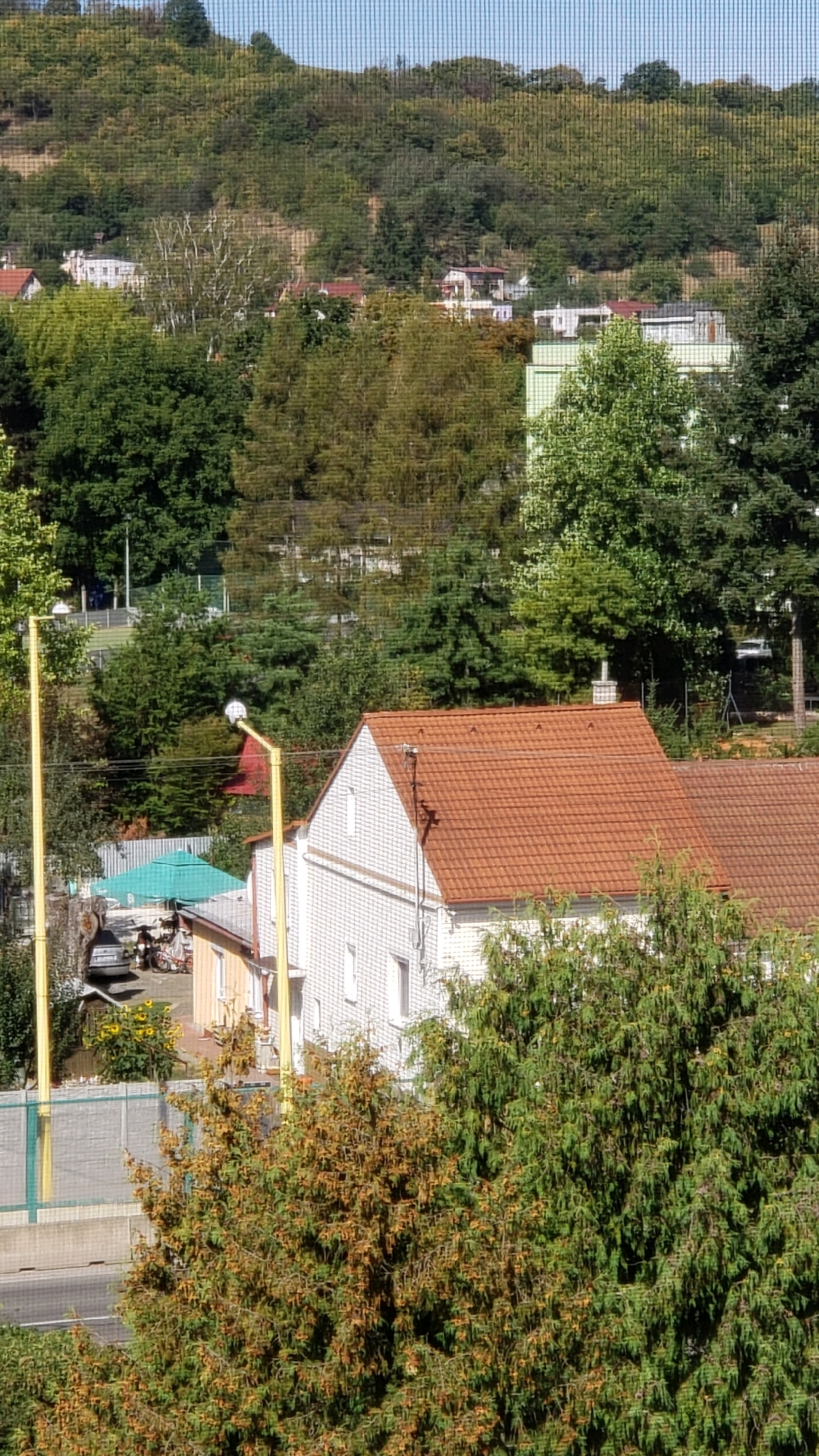 The image captures a suburban residential area on a hillside in Prešov, Slovakia. The foreground is dominated by dense green foliage, with some trees showing autumnal colors. A two-story house with a red tiled roof and white brick exterior is prominently featured. To the left of this house, a smaller building with a lighter colored facade and a light-colored roof is visible. Adjacent to this smaller building is a grey concrete fence. Further to the left, a yellow pole with a light fixture stands next to a white and grey fence. Beyond these immediate structures, a network of trees covers the hillside, with several other residential buildings scattered amongst them. The background consists of a densely forested hill under a clear blue sky. A road is visible in the lower part of the image, running horizontally.