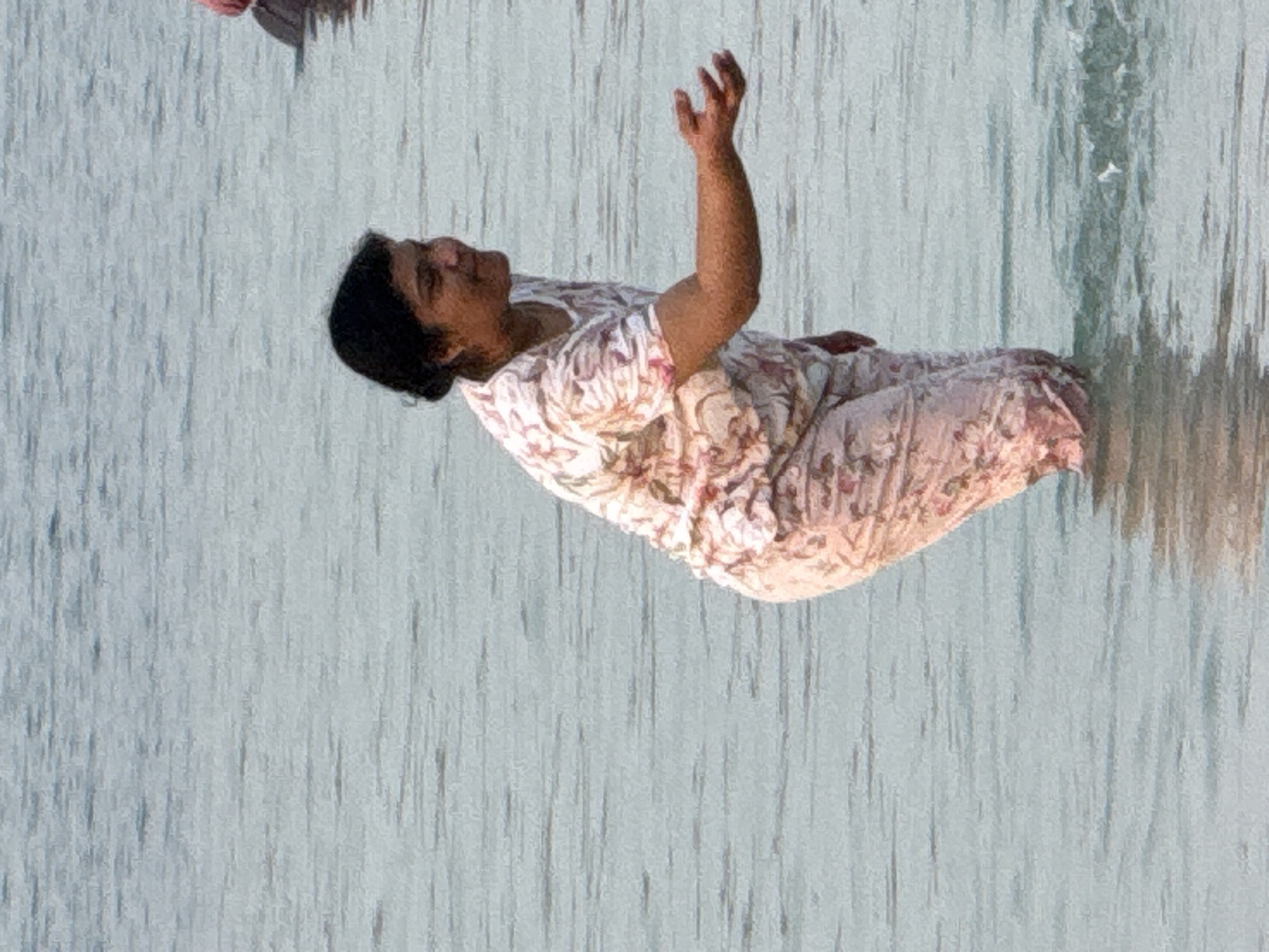 A woman wearing a floral print shirt and pants stands in shallow, calm water. Her hair is pulled back in a bun. She is facing to her right, with her left arm extended forward, palm open, as if reaching out to someone or something. The water is a pale blue-gray and appears to be a body of water such as a lake, river, or the sea. The background is a blurred expanse of water. The lighting suggests it is daytime. The location is not specified in the image or accompanying data.