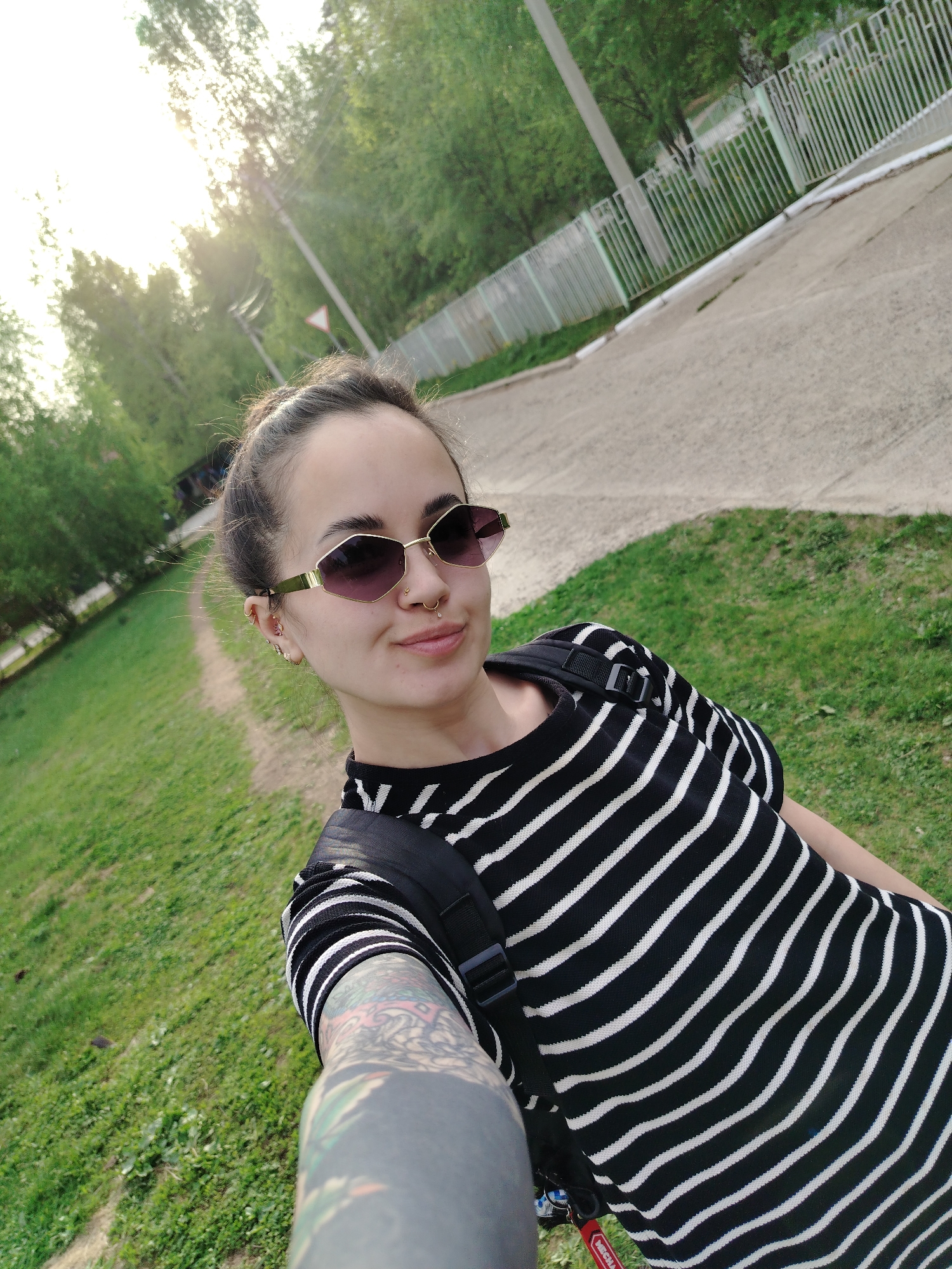A young woman stands outdoors, taking a selfie. She has her hair pulled back in a bun, and is wearing sunglasses, a black and white striped shirt, and a backpack. Visible on her arm is a tattoo. In the background, there is green grass, a path, trees, a road, and a white fence. The location is Noviy, Russia.