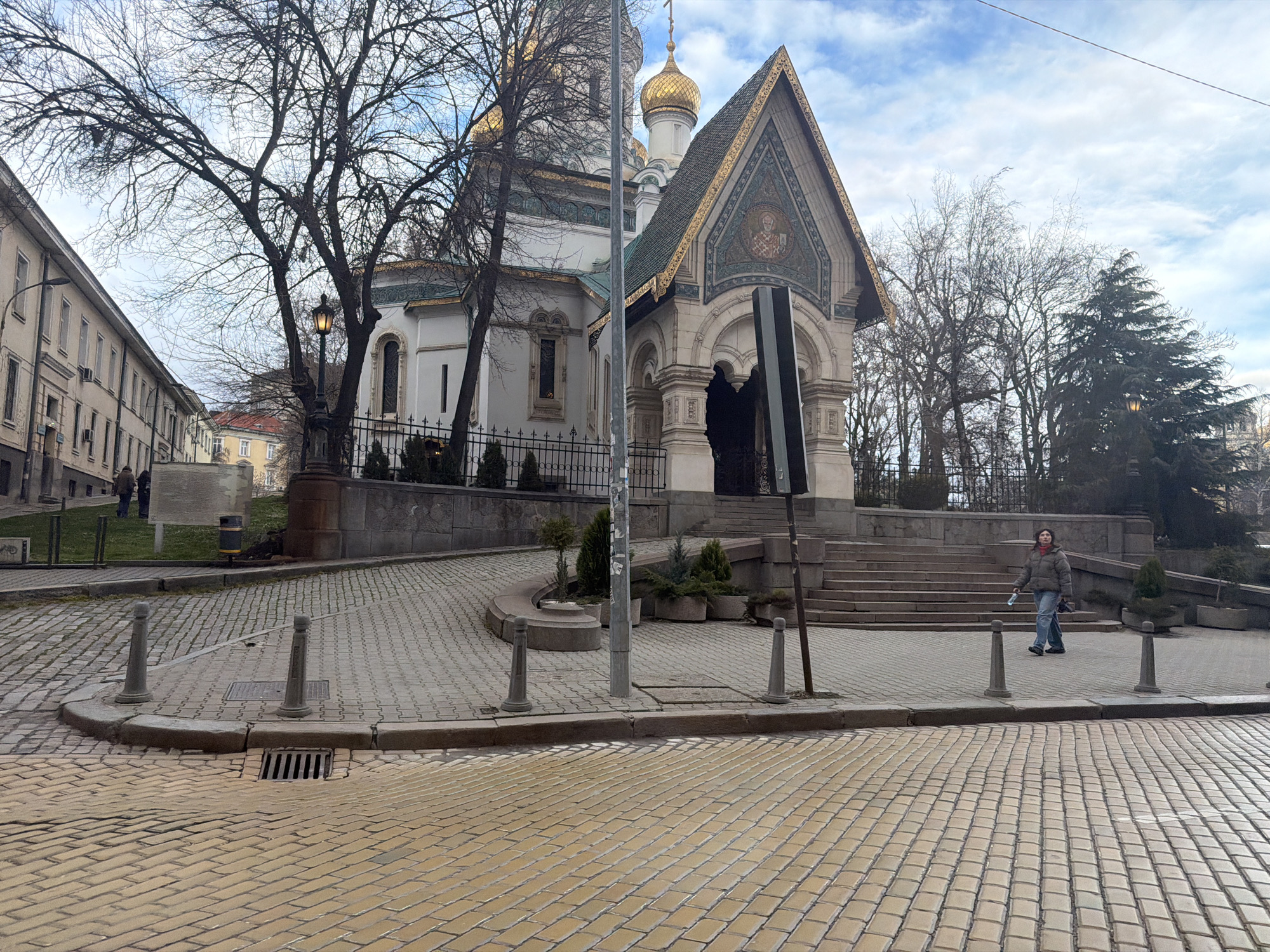 The image depicts a street scene in Sofia, Bulgaria, specifically near the St. Alexander Nevsky Cathedral and St. George Chapel. The foreground shows a cobblestone street intersection. A woman in a green jacket and jeans is walking towards the camera on the sidewalk, which is also paved with cobblestones. There are stone bollards marking the edge of the sidewalk. A streetlamp and various bare trees are visible. The St. George Chapel, with its golden domes and intricate mosaic detailing, is a prominent feature of the image, set back from the street and slightly elevated. Part of the Alexander Nevsky Cathedral is also visible behind the chapel. A fence runs along the perimeter of the chapel grounds. The sky is overcast with patches of blue. Several buildings are visible in the background to the left.