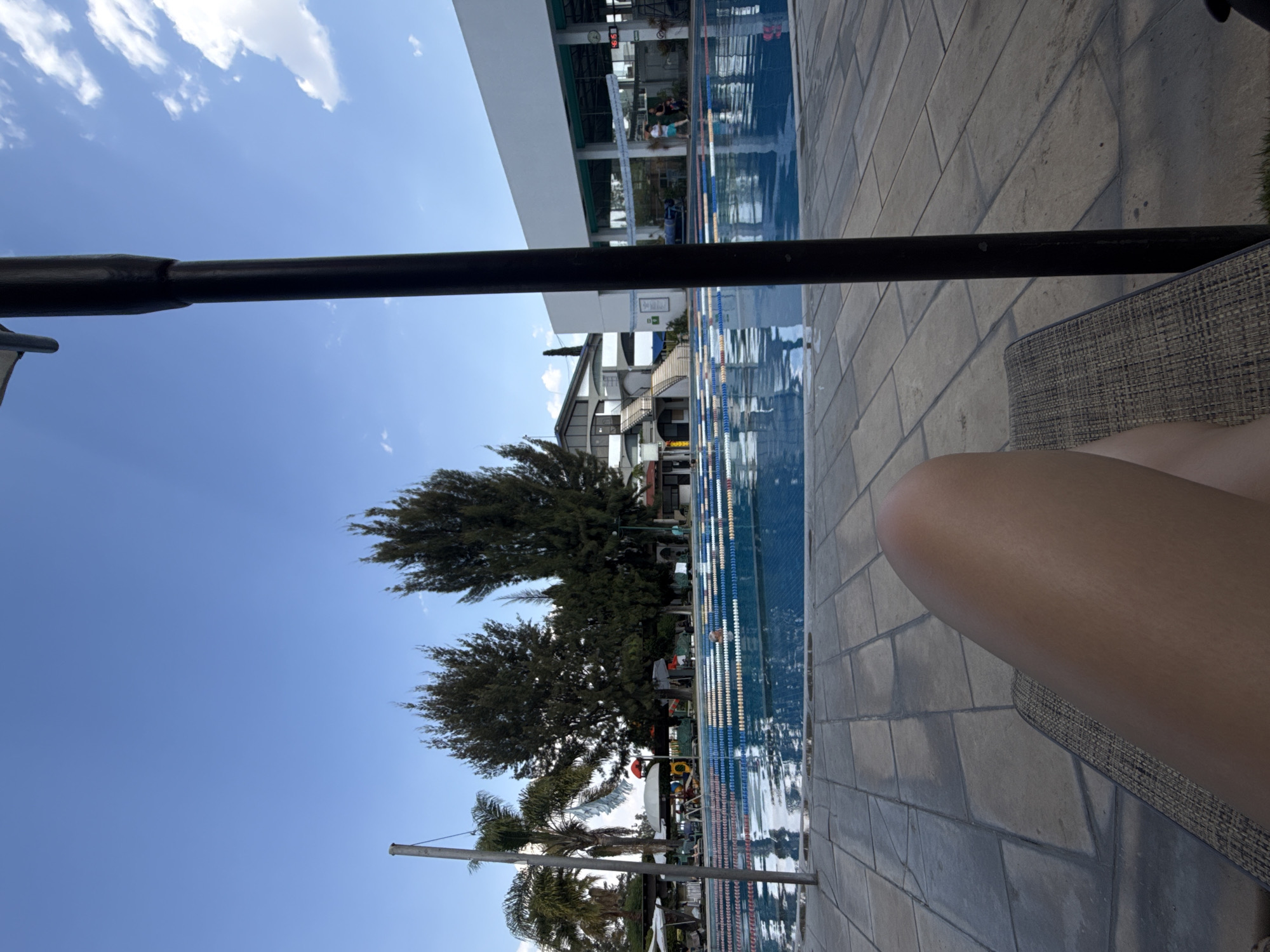 The image captures a scene at a swimming pool complex. The foreground shows a portion of a person's thigh resting on a woven-texture lounge chair. In the midground, there's an outdoor swimming pool with multiple lanes, marked by lane dividers. People are visible in and around the pool. Palm trees and tall, broad-leafed trees line one side of the pool area. Behind the trees is a building with multiple stories. The sky is blue with scattered clouds. An angled black metal bar partially obstructs the view.