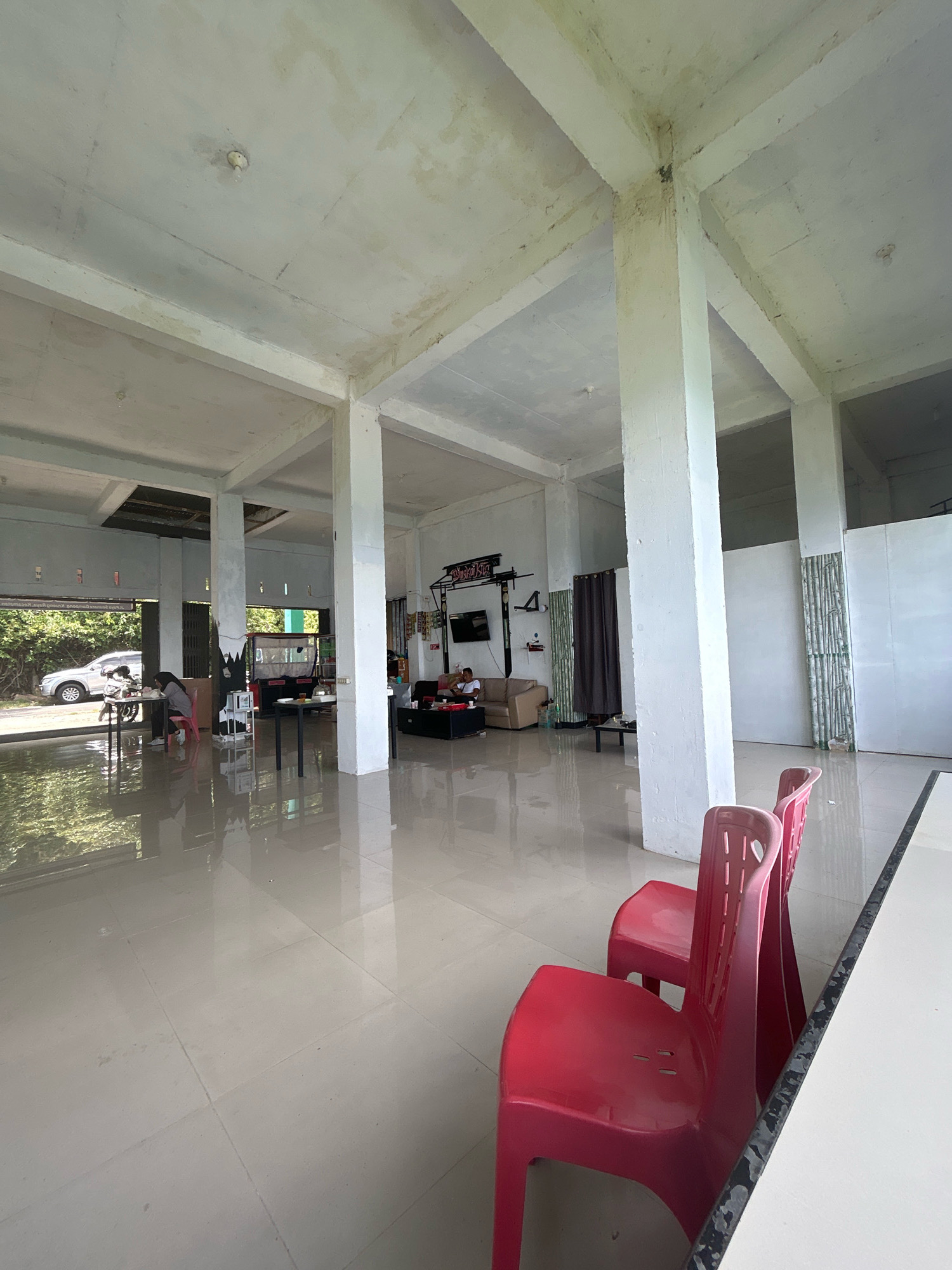 The image shows the interior of a simple, open-air restaurant or cafe. The space has a concrete floor, columns, and ceiling, with natural light entering from the open front. In the foreground, two bright red plastic chairs are visible. Further back, several tables are set up, with some people seated and interacting. One person is at a table near the entrance, and two others are seated on a sofa in a lounge area. The background shows an outdoor view with trees and a parked car. The overall impression is of a casual, functional establishment in a tropical setting, likely Sabang, Indonesia, given the architectural style and climate.