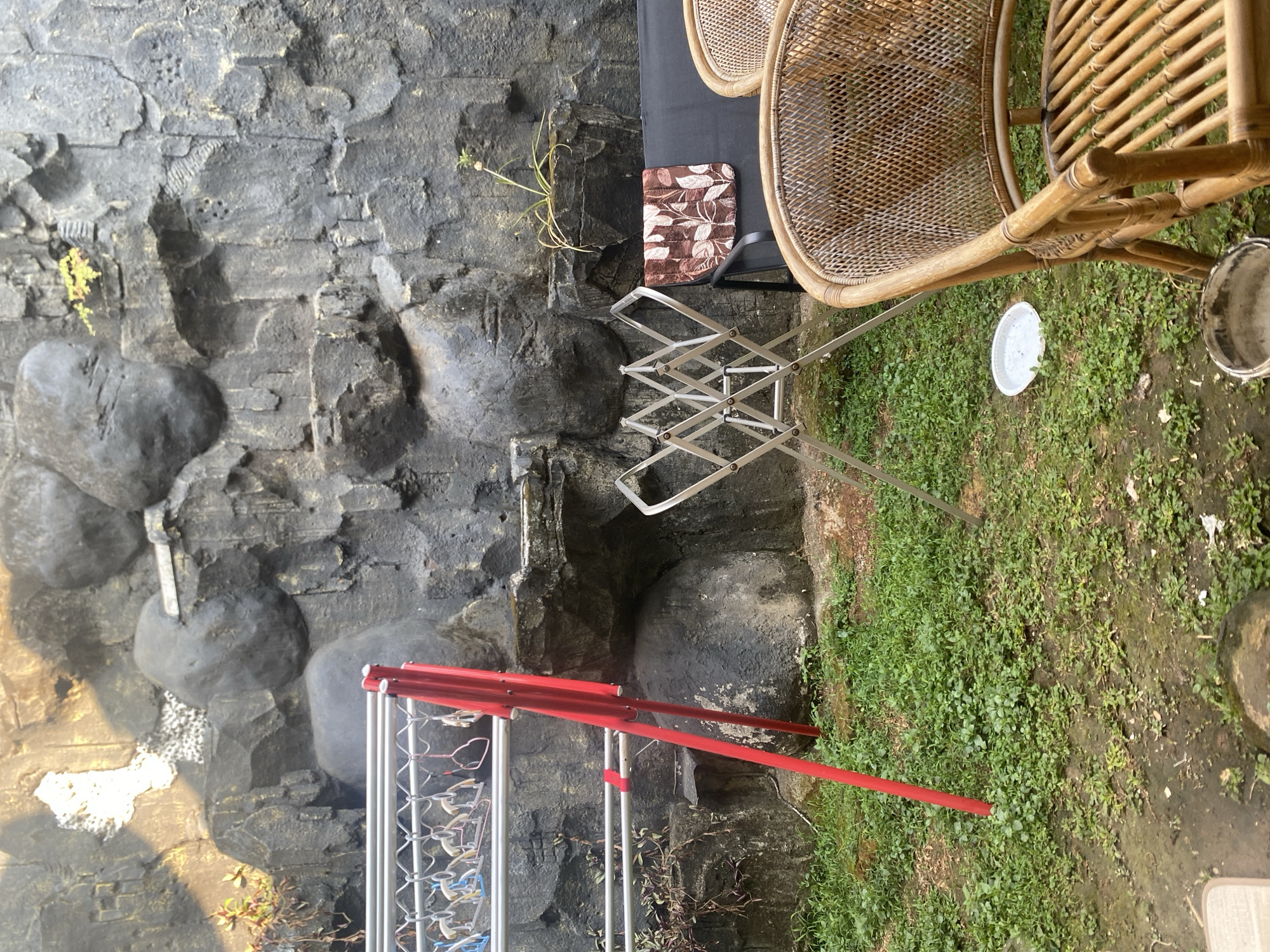 In Dukuh, Indonesia, an outdoor scene unfolds against a backdrop of a textured, dark gray rock wall, possibly artificial. In the foreground, lush green grass covers the uneven ground. To the left, a red metal clothes drying rack with several hangers is set up. To the right of the drying rack, a collapsible silver drying rack stands open. Further to the right, a dark-colored sofa or bench is visible, draped with a brown and white patterned fabric. Two wicker chairs, with intricate woven backs and seats, are positioned in the immediate right foreground. A small white plastic plate rests on the grass near the silver drying rack. In the background, the rock wall features varying textures and some small green plants emerge from crevices. The lighting suggests it is daytime, with some sunlight casting shadows. There are no people visible in the scene.