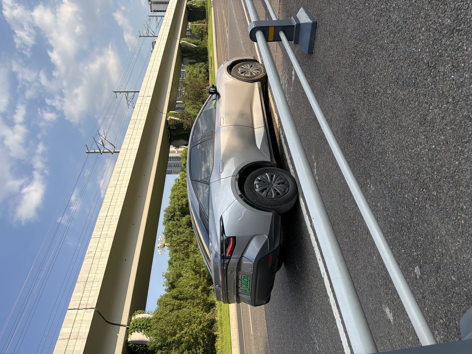 A silver Toyota bZ4X electric vehicle, license plate A·C00256, is parked on a paved road in Jiangjin District, Chongqing, China. The car is viewed from the rear-left, with its taillights illuminated. Behind the car, a concrete overpass with visible structural supports overgrown with ivy stretches across the frame. Overhead, electrical lines associated with a train or monorail system are suspended. The sky is blue with scattered white clouds, and a line of trees and buildings are visible in the background. The foreground features a metal guardrail with dark gray posts and light gray horizontal bars, positioned along the edge of the road.