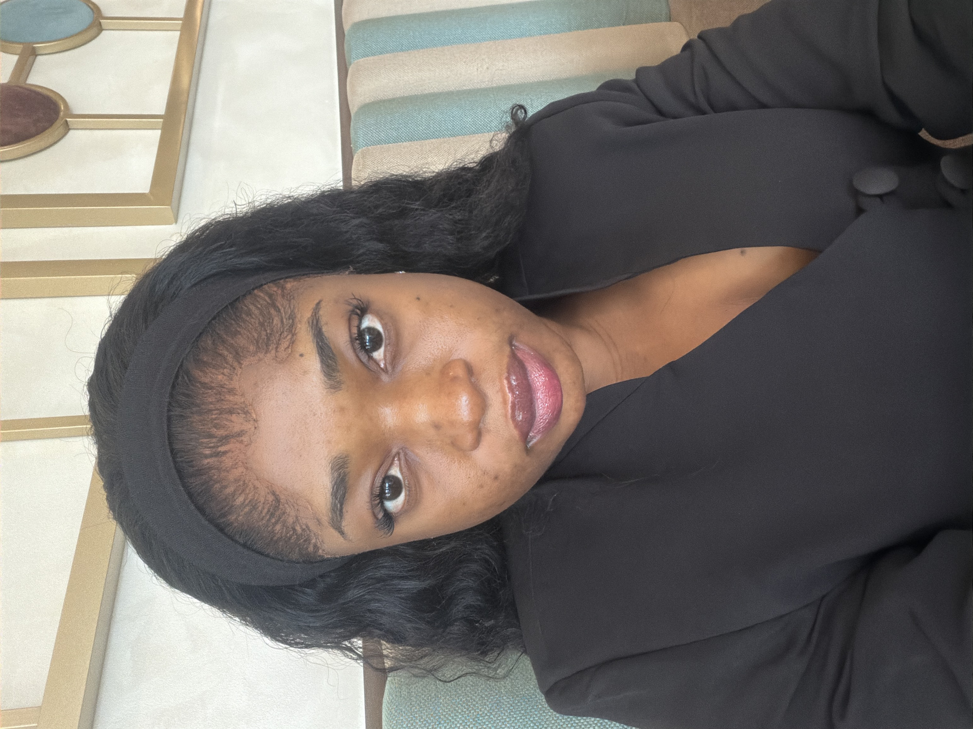 A woman with dark hair and a black headband is wearing a black blazer. The background shows a wall with a decorative gold-framed pattern and a striped cushion or seating area in light blues, beige, and white. The location in Abuja, Nigeria, cannot be determined from the image provided.