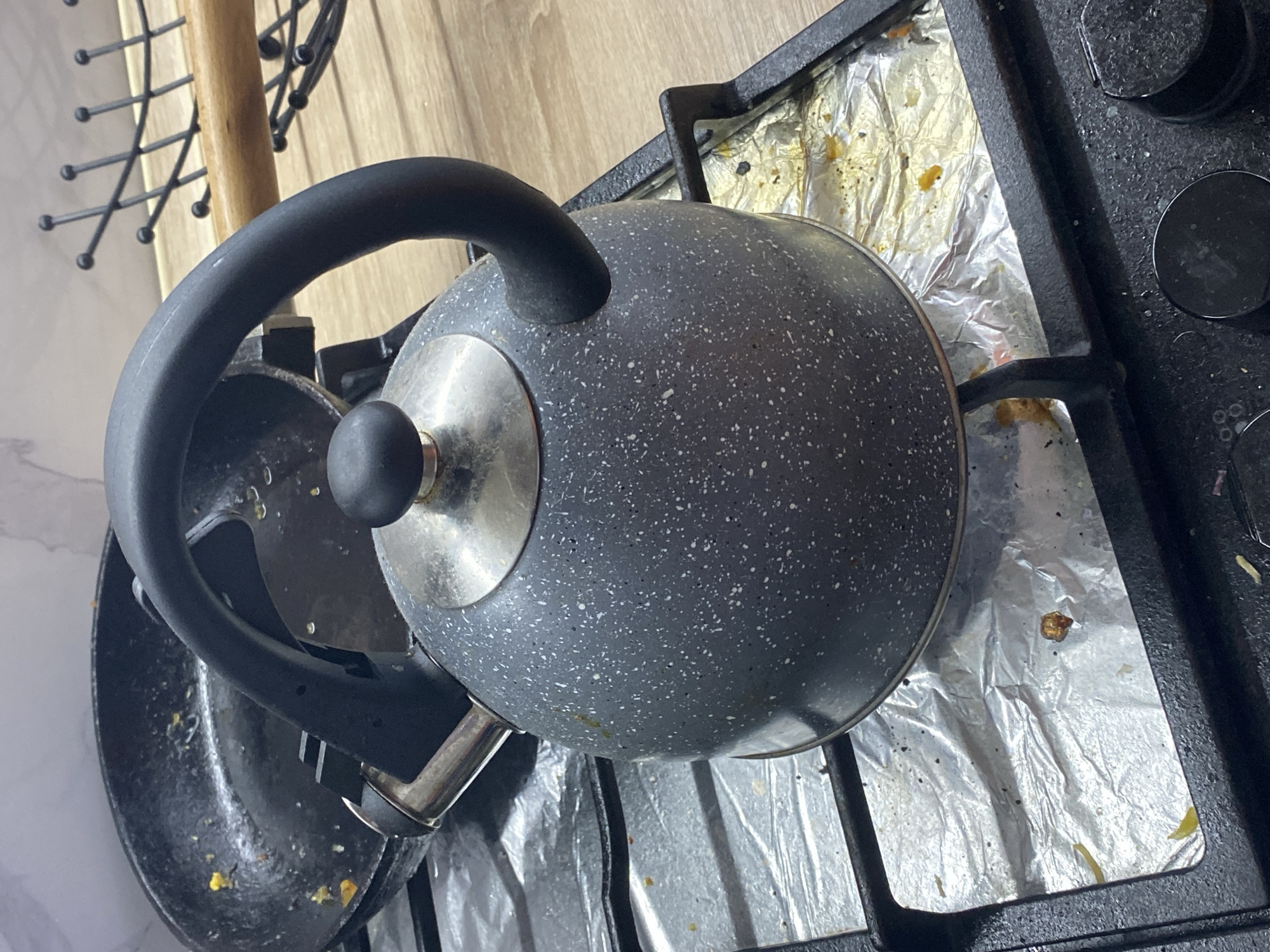 A speckled gray kettle with a black handle sits on a gas stove. The stove burner beneath the kettle is covered with crumpled aluminum foil, which appears stained with food residue. To the left of the kettle is a black frying pan, also with visible food debris inside. The background shows a wooden countertop and a black metal rack with wooden tongs. The scene appears to be a domestic kitchen environment. The image does not provide information about the city of Kryvyi Rih, Ukraine.