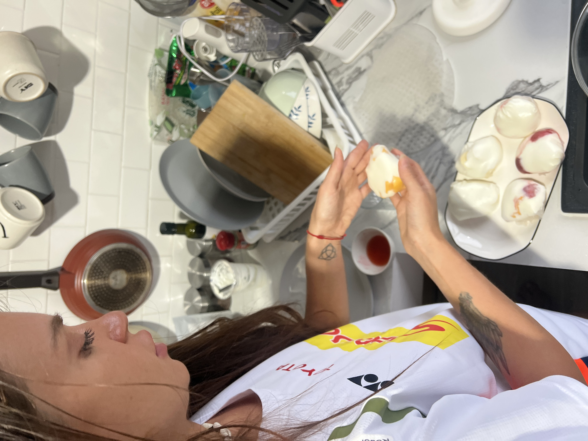 The image depicts a person in a white and red sports shirt holding a peeled egg in their hands. In the background, there is a white countertop filled with various kitchen items including cups, bowls, a frying pan, a cutting board, bottles, and a dish rack. On a plate next to the person's hands are several other peeled eggs. The location is unclear, but presumably in a kitchen. There is no visible indication of บ้านบางรักษ์, Thailand.