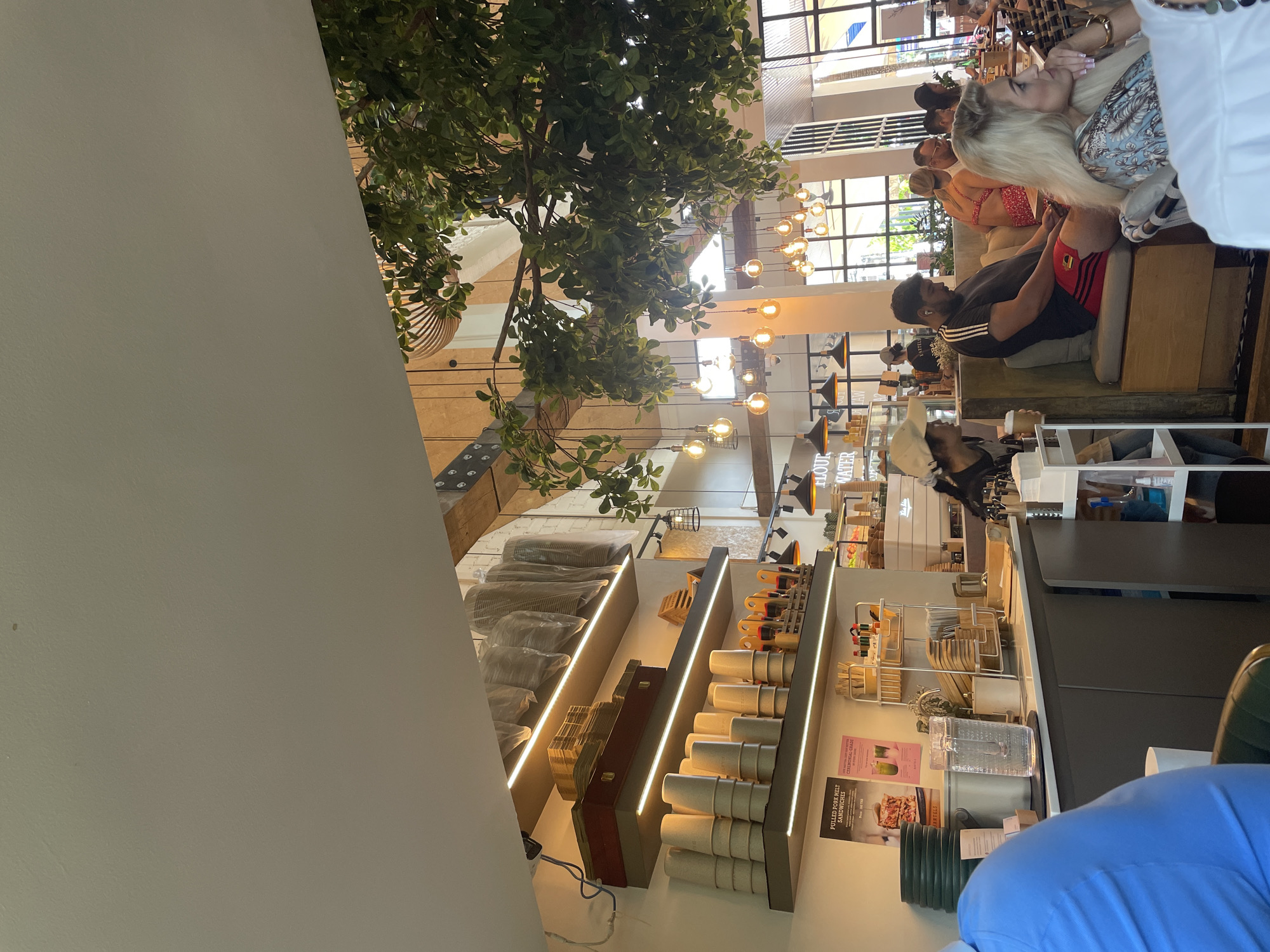 The image captures an interior scene, likely a café or restaurant, in Choeng Thale, Thailand. Tall windows let in light. Hanging light fixtures illuminate the space. Several people are seated on a cushioned bench, and they appear to be engaged in conversation. One of them has fair hair, and another is wearing a black shirt with red detailing. A display of cups and takeout boxes stands near the counter. Above are two signs that say "Flour Water". A green, leafy tree sits near the white curved ceiling.