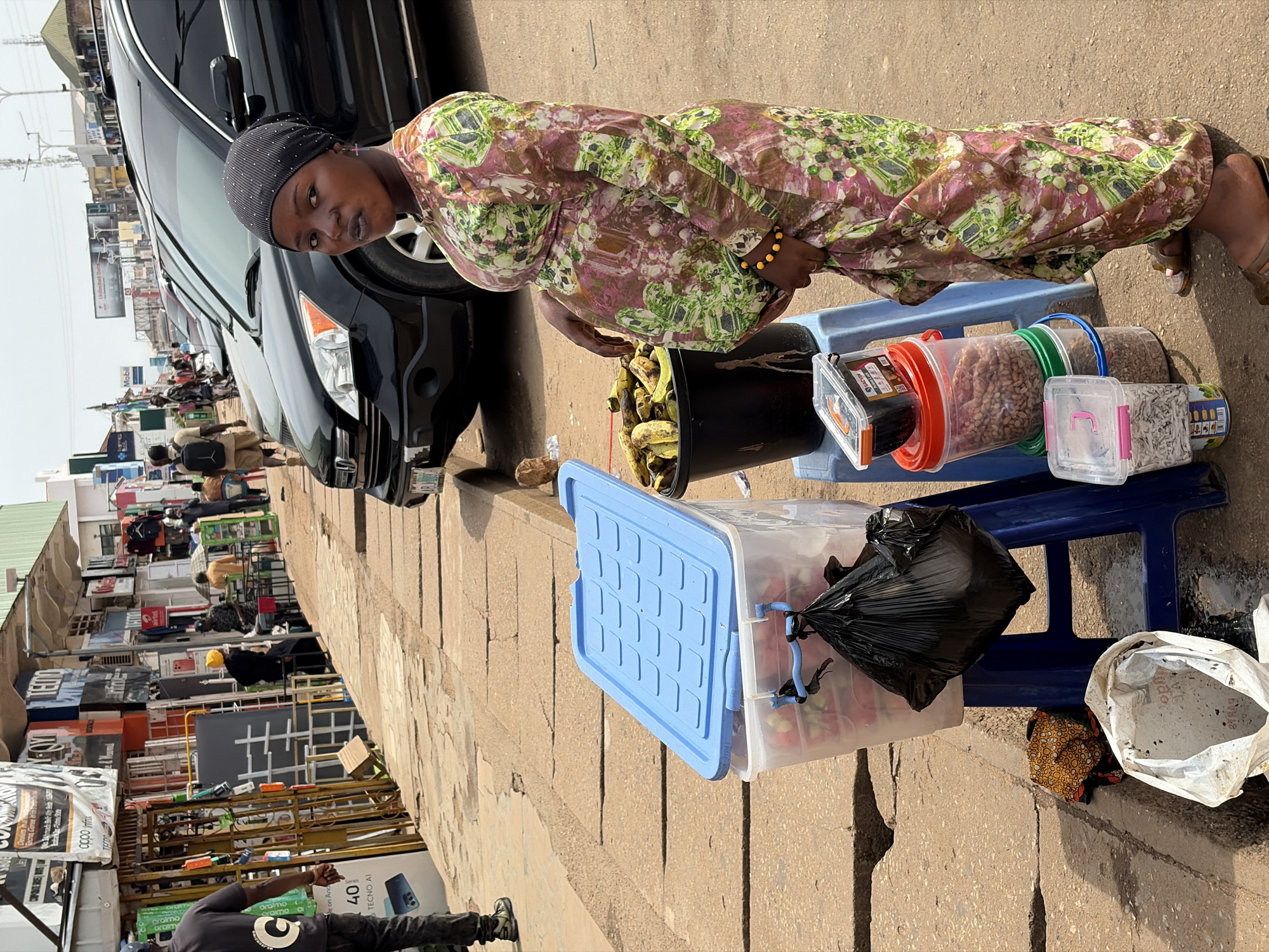In Jos, Nigeria, a pregnant woman stands on a sidewalk next to a street market. She is wearing a colorful floral dress and a headscarf. In front of her, there are several containers of goods, including a large blue bin with a lid, a bucket of bananas, and various plastic containers filled with food items. A black plastic bag hangs from one of the containers. In the background, there are shops and cars parked along the street. People can be seen walking and shopping in the market. The environment is a bustling urban street scene.
