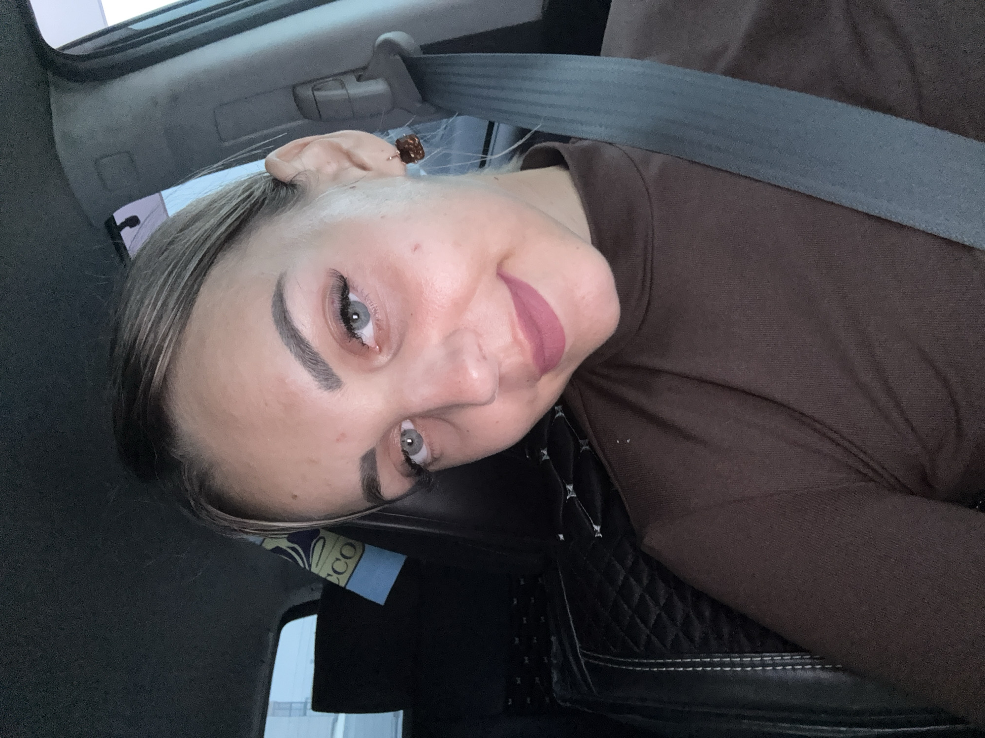 A young woman is taking a selfie in the passenger seat of a car. She is wearing a brown turtleneck shirt and a seatbelt. Her hair is pulled back, and she is wearing makeup, including eyeliner and lipstick. She has light blue eyes and fair skin. The background shows the interior of the car, including a headrest with a patterned cover and a seatbelt buckle. There is also a piece of fabric with the word "ECCO" visible in the background. The lighting suggests it is nighttime or a very dimly lit environment. The image was taken in Yuzhno-Sakhalinsk, Russia.