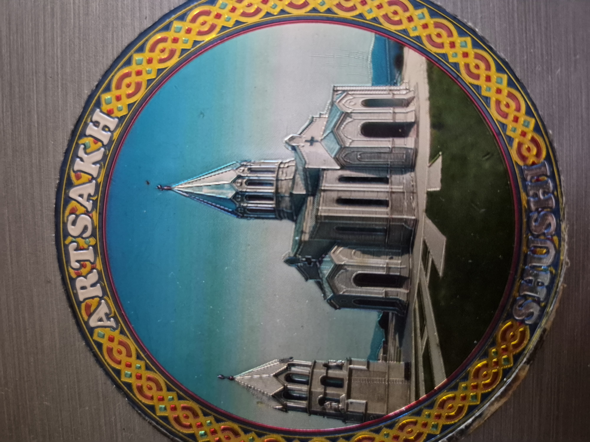 This media file displays a circular commemorative item, likely a magnet or badge, featuring a prominent architectural landmark. The central image, oriented sideways, depicts a grand, light-colored building with intricate details, multiple towers, and pointed spires, set against a clear blue sky and some green ground. The building's design suggests a religious structure, specifically a cathedral, with visible crosses on its spires.
The circular image is framed by a decorative border featuring a repeating pattern of yellow, red, and blue geometric motifs. Along this border, white capital letters spell out two distinct names. On the left side, reading vertically from bottom to top (when viewed upright), the word "ARTSAKH" is visible. On the right side, reading vertically from top to bottom (when viewed upright), the word "SHUSHI" is inscribed.
Given the text, the building depicted is the Ghazanchetsots Cathedral (Holy Savior Cathedral) located in Shushi, a city within the Artsakh Republic, which holds significant cultural importance to Armenians. The clear sky suggests a daytime scene. No people or specific activities are shown; the image is a static representation of the landmark. The edges of the item show slight signs of wear. The context of Yerevan, Armenia, aligns with the cultural significance of Artsakh and Shushi to Armenians.