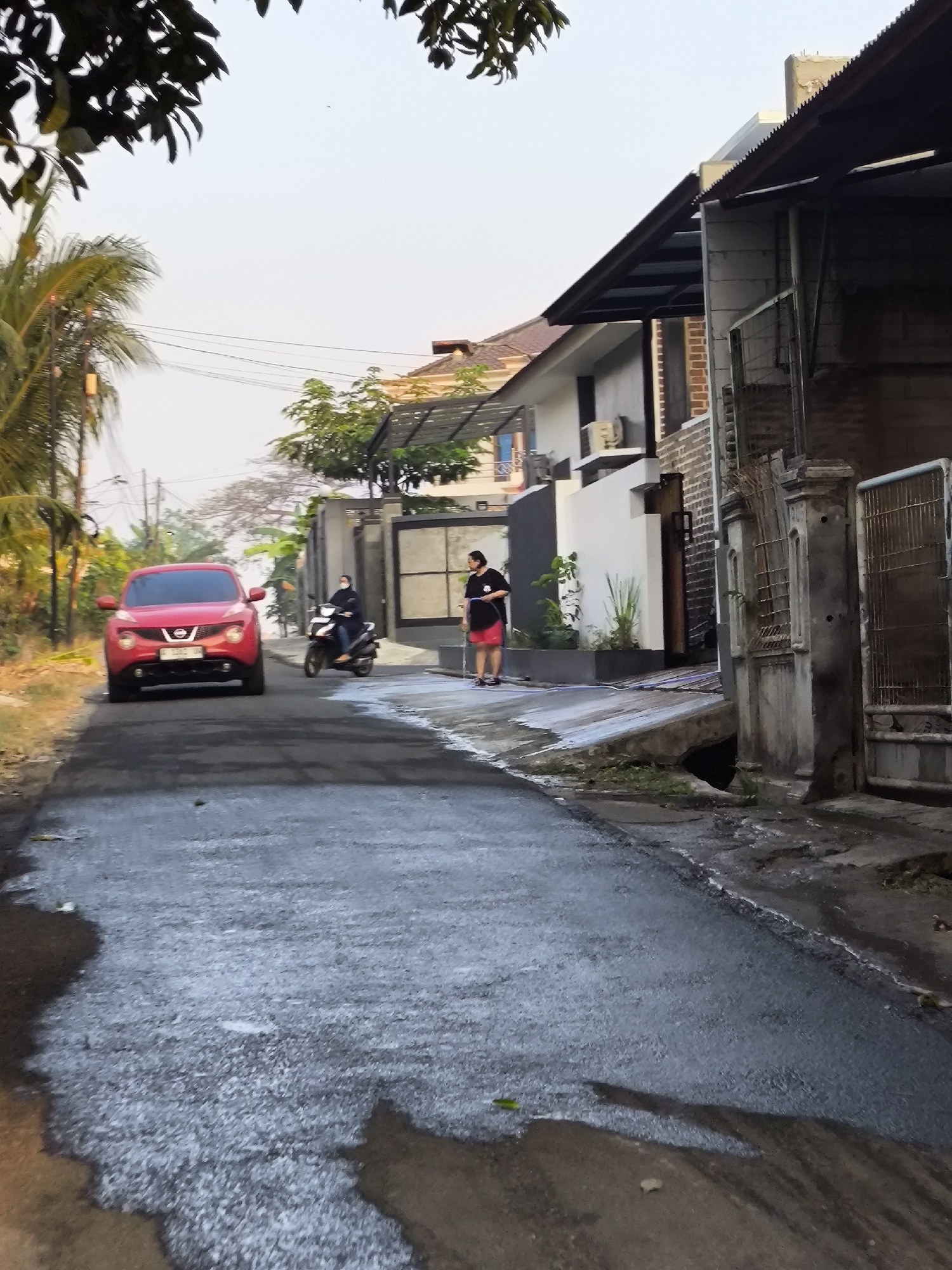 A red Nissan Juke is parked on an asphalt road in a residential area in Cilegon, Indonesia. The car is facing the camera, and its headlights are on. A person is visible riding a scooter in the background, heading away from the camera. Another person is standing on the side of the road near a gate. The road appears wet, suggesting recent rain. Buildings with modern architecture line the street, and some trees and foliage are visible. The sky is overcast.