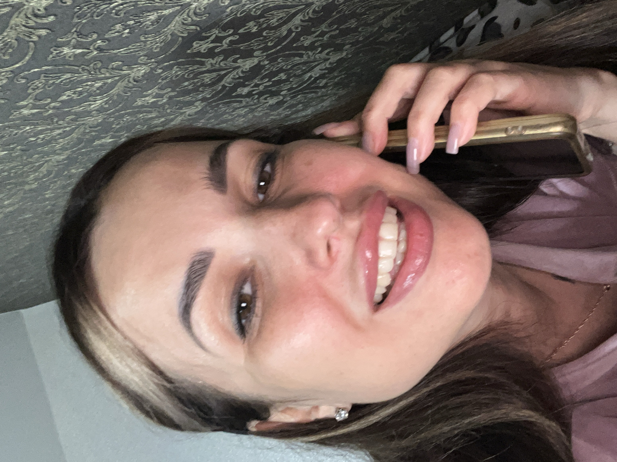 A close-up selfie shows a woman holding a gold-colored smartphone to her right ear, looking directly at the camera. She has long, dark brown hair with blonde highlights, fair skin with a reddish blush on her cheeks, dark eyebrows, and brown eyes. Her lips are painted with a glossy pink lipstick, and she is smiling, revealing white teeth. She is wearing a pink top and a delicate gold necklace with a small pendant. A diamond stud earring is visible in her left ear. The background on the right is a dark wallpaper with a damask pattern, while the left side shows a light blue wall. The scene appears to be indoors.